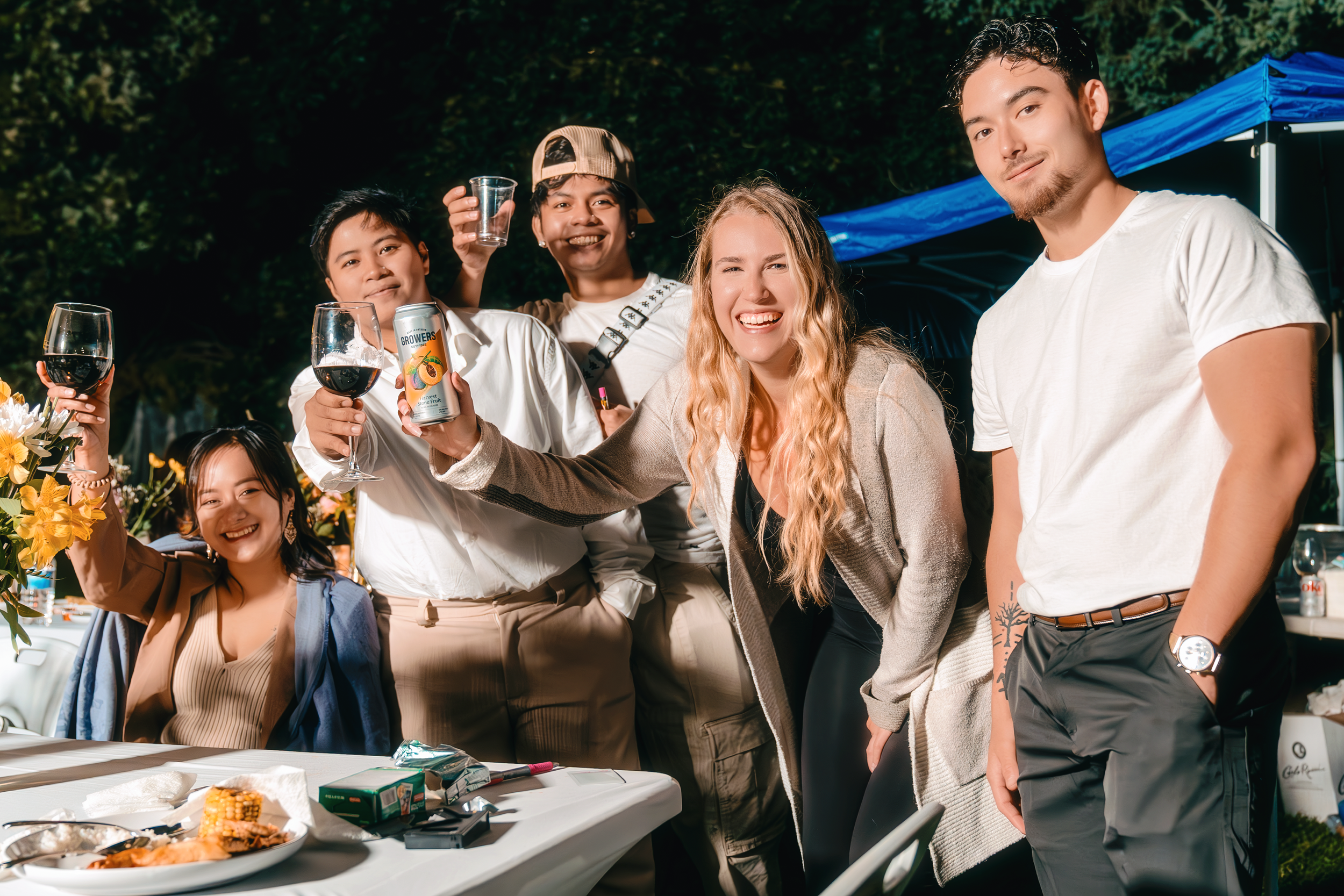 Group of friends celebrating at an outdoor party with glasses of wine and food on the table, smiling and enjoying the evening.