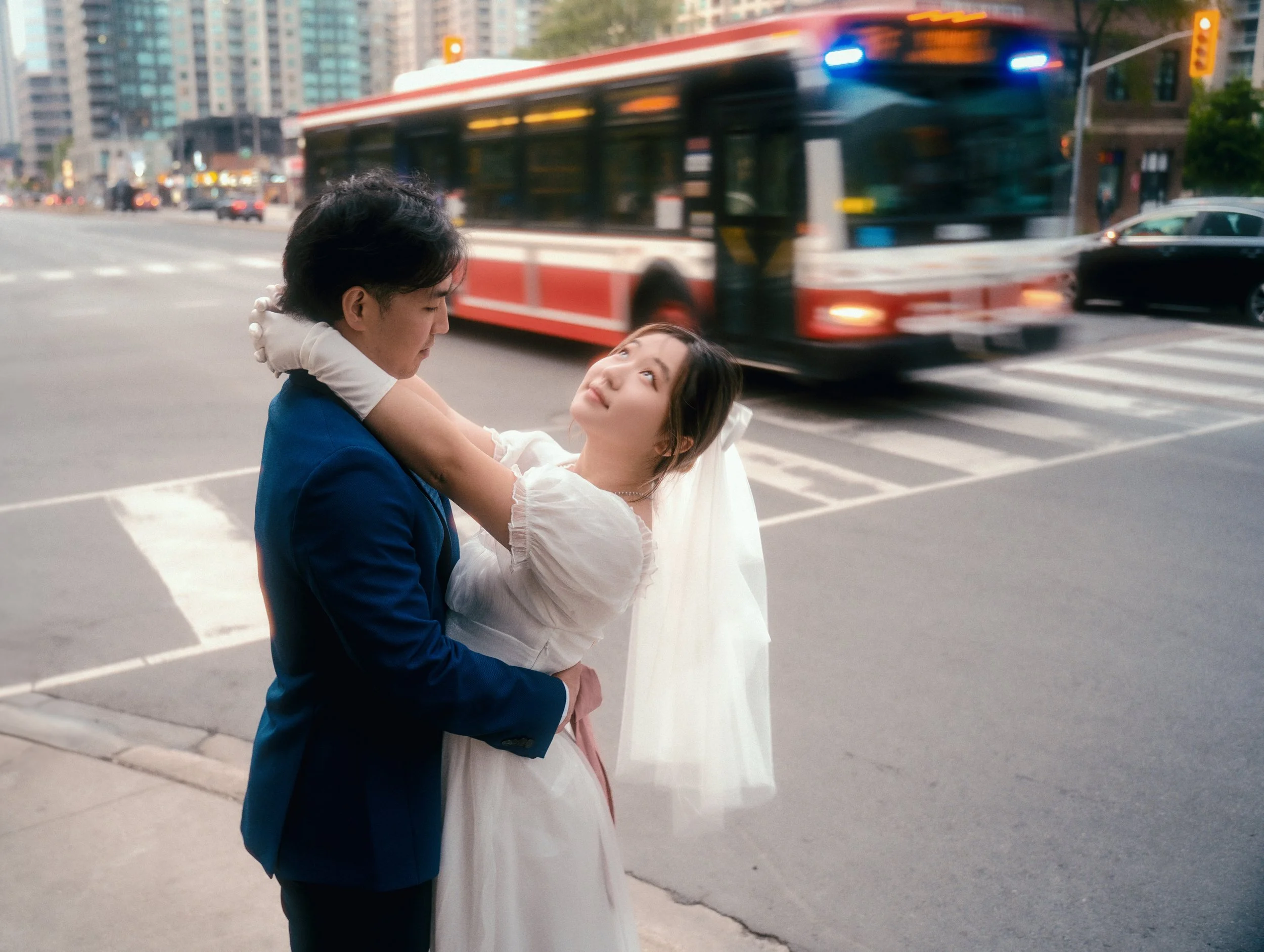 A young couple dressed in wedding attire on a city street, with the groom in a blue suit and the bride in a white dress and veil, sharing an intimate moment as a red and white bus passes by in the background.