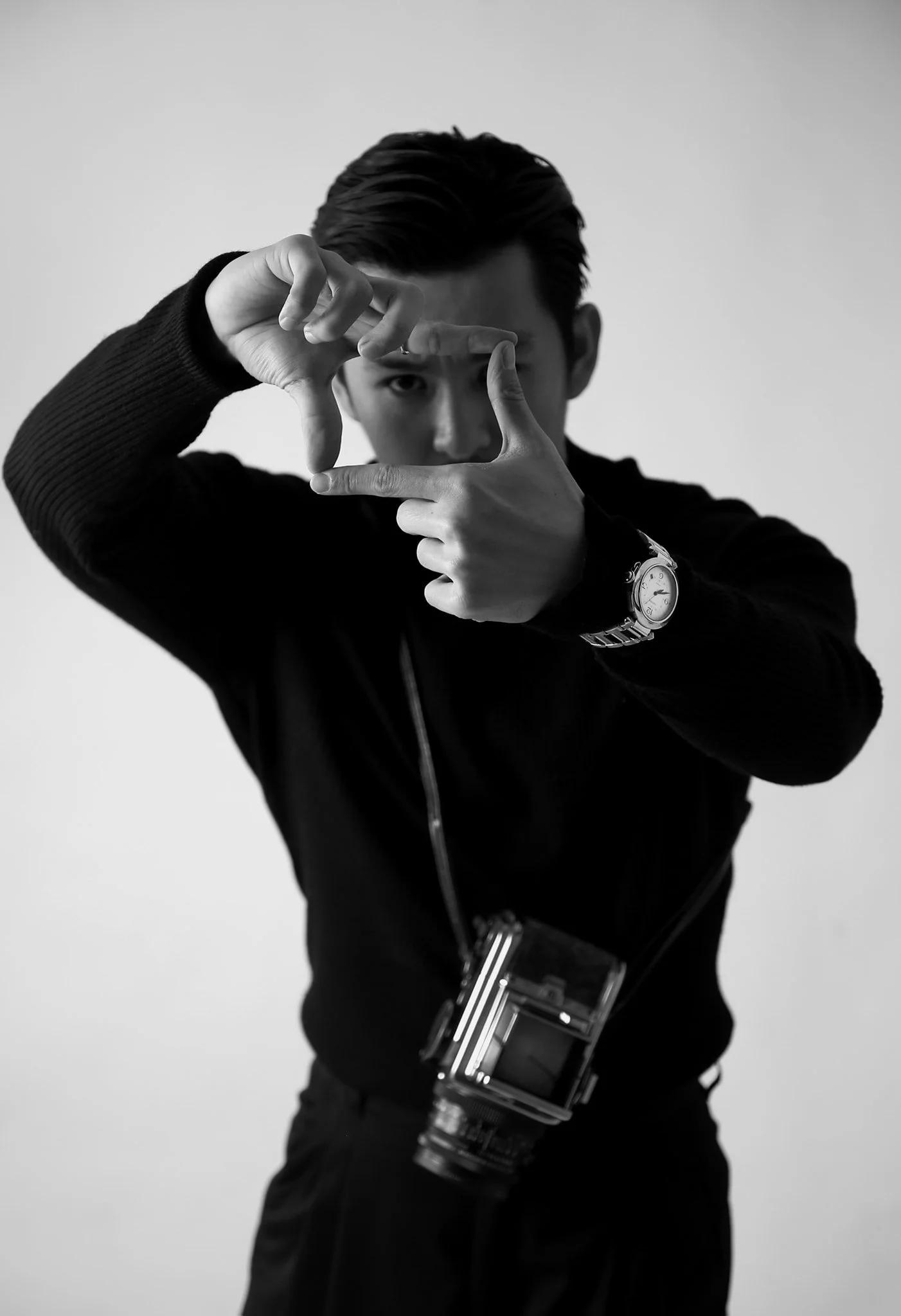 A young man with dark hair, wearing a black sweater and a wristwatch, is making a framing gesture with his hands and looking through an imaginary viewfinder. He has a camera hanging around his neck and is standing against a plain light background.