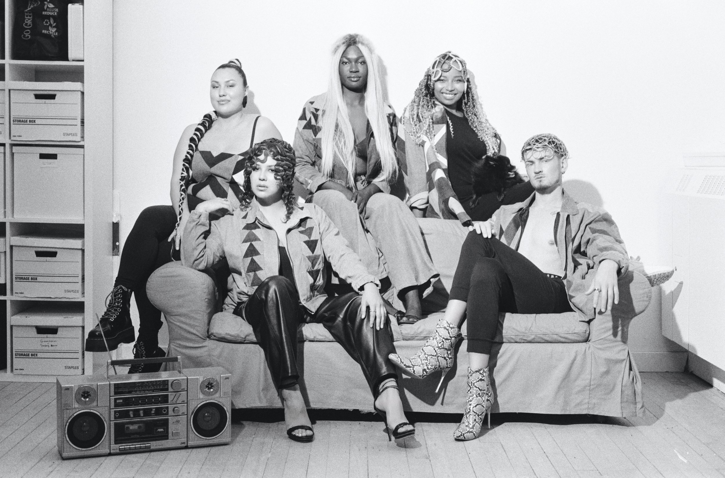 Six young adults posing on a couch and around it in a room with a stereo, cardboard storage boxes, and a plain wall.