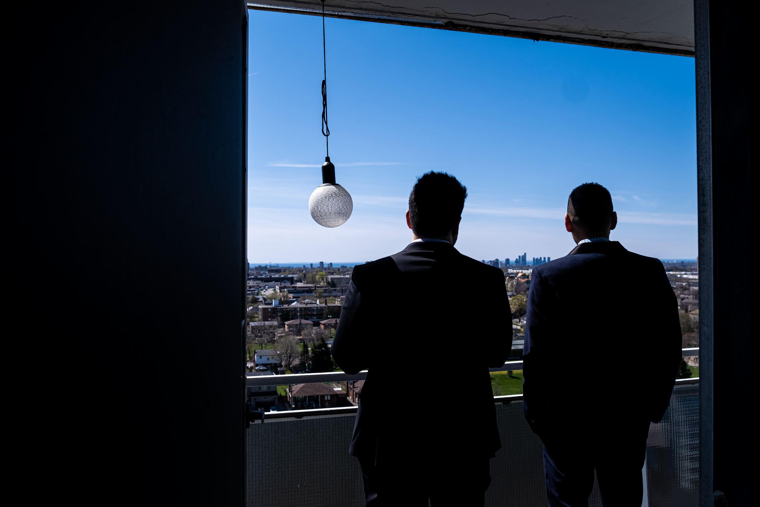 Two men in suits standing on a balcony, looking out at a city skyline on a clear day.