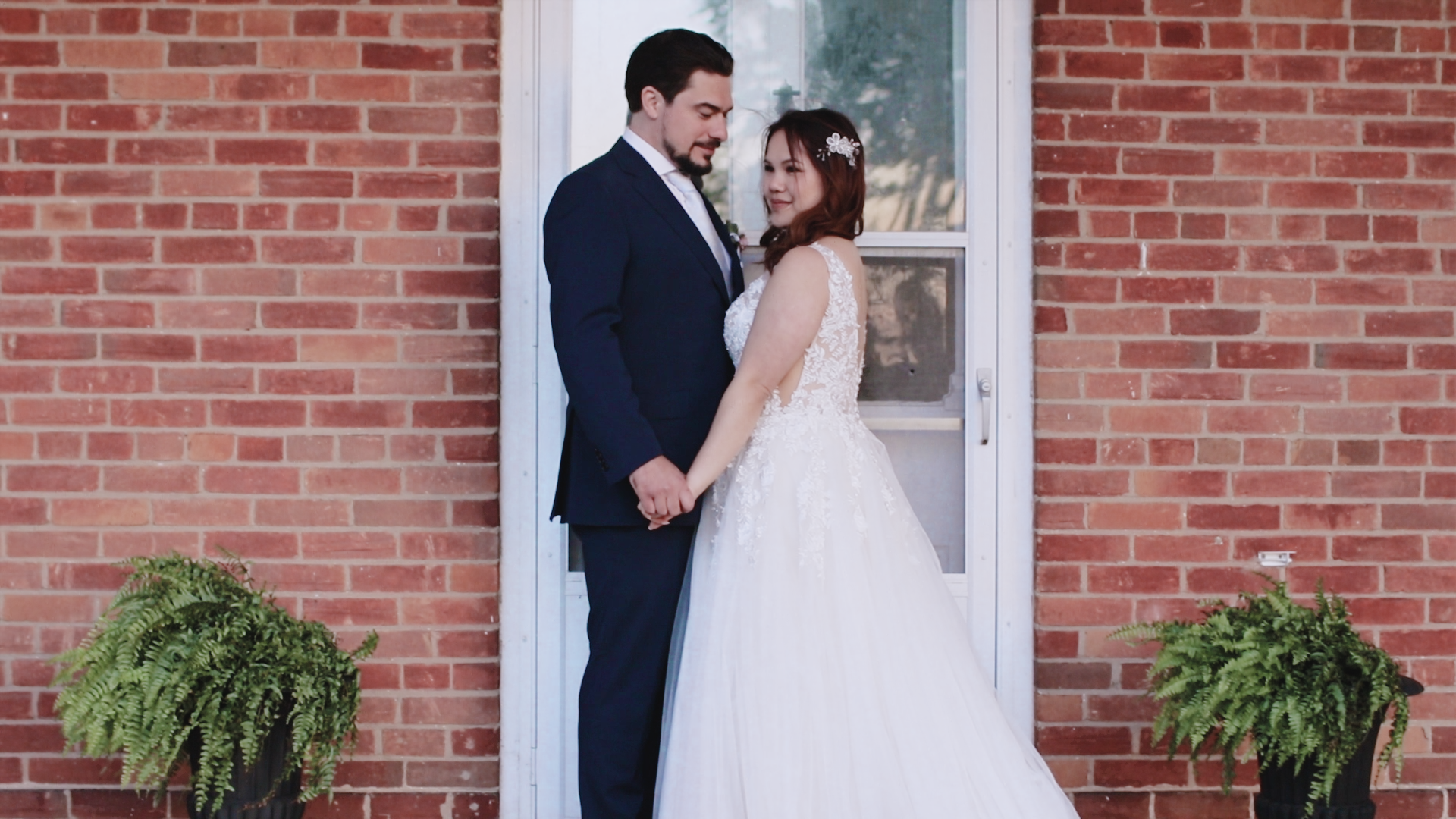 A bride and groom standing hand in hand outside a brick building, with greenery in pots on either side of them, during their wedding.