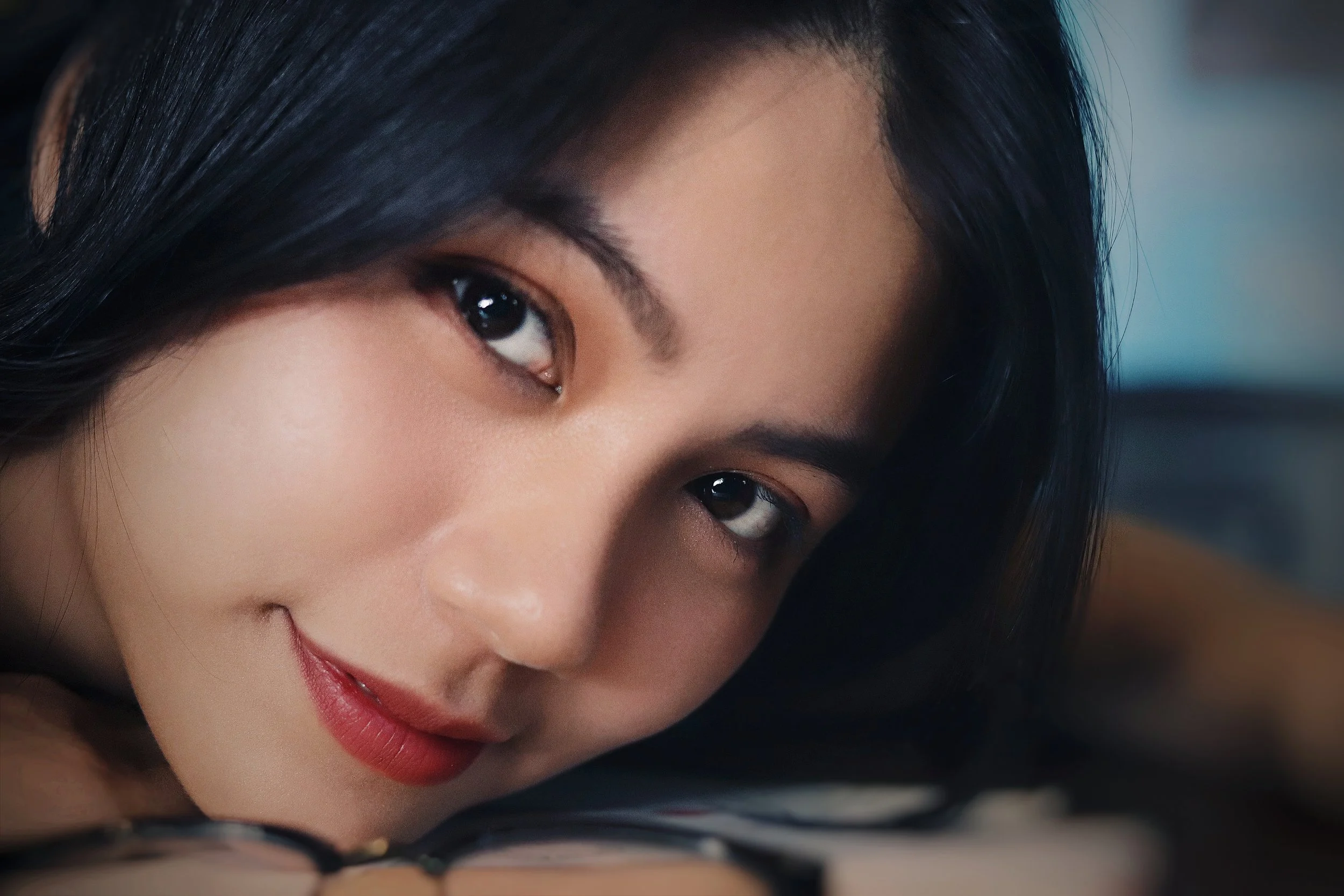 Close-up of a woman with dark hair, lying down with her face touching a surface, smiling softly and looking at the camera.