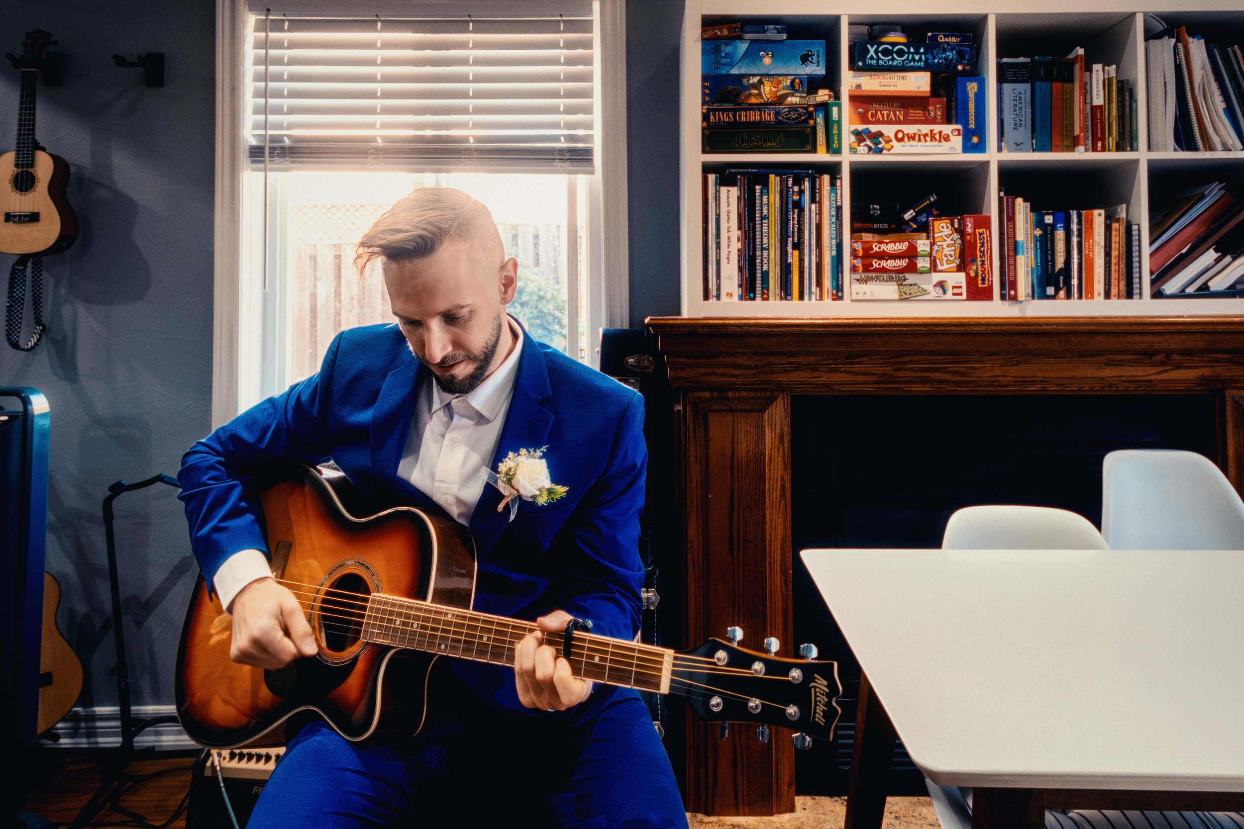 A man in a blue suit playing an acoustic guitar indoors, with bookshelves in the background and sunlight coming through a window.