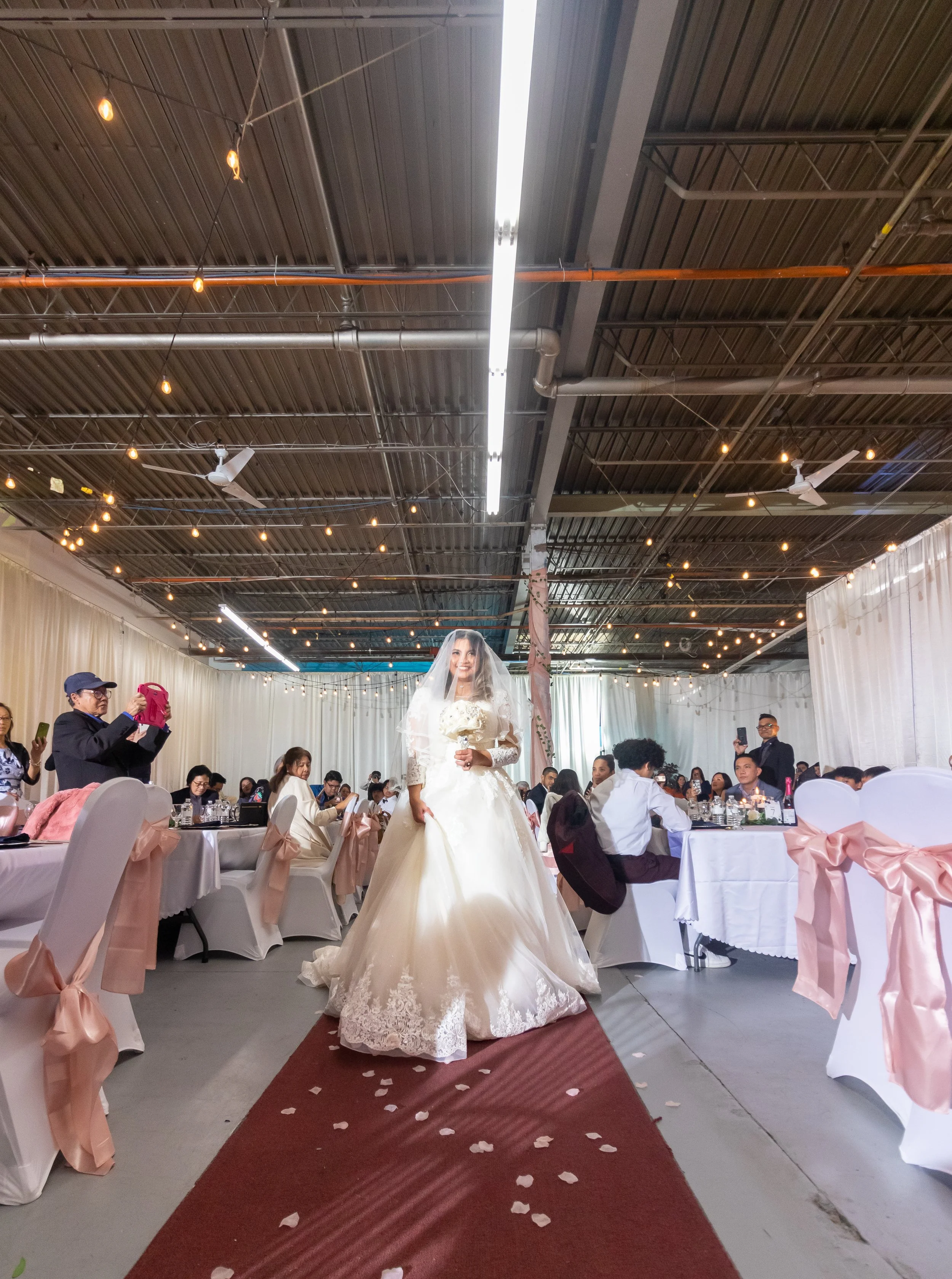 Bride in a white wedding dress and veil holding a bouquet of white flowers walking down the aisle at a wedding reception with guests seated at decorated tables.