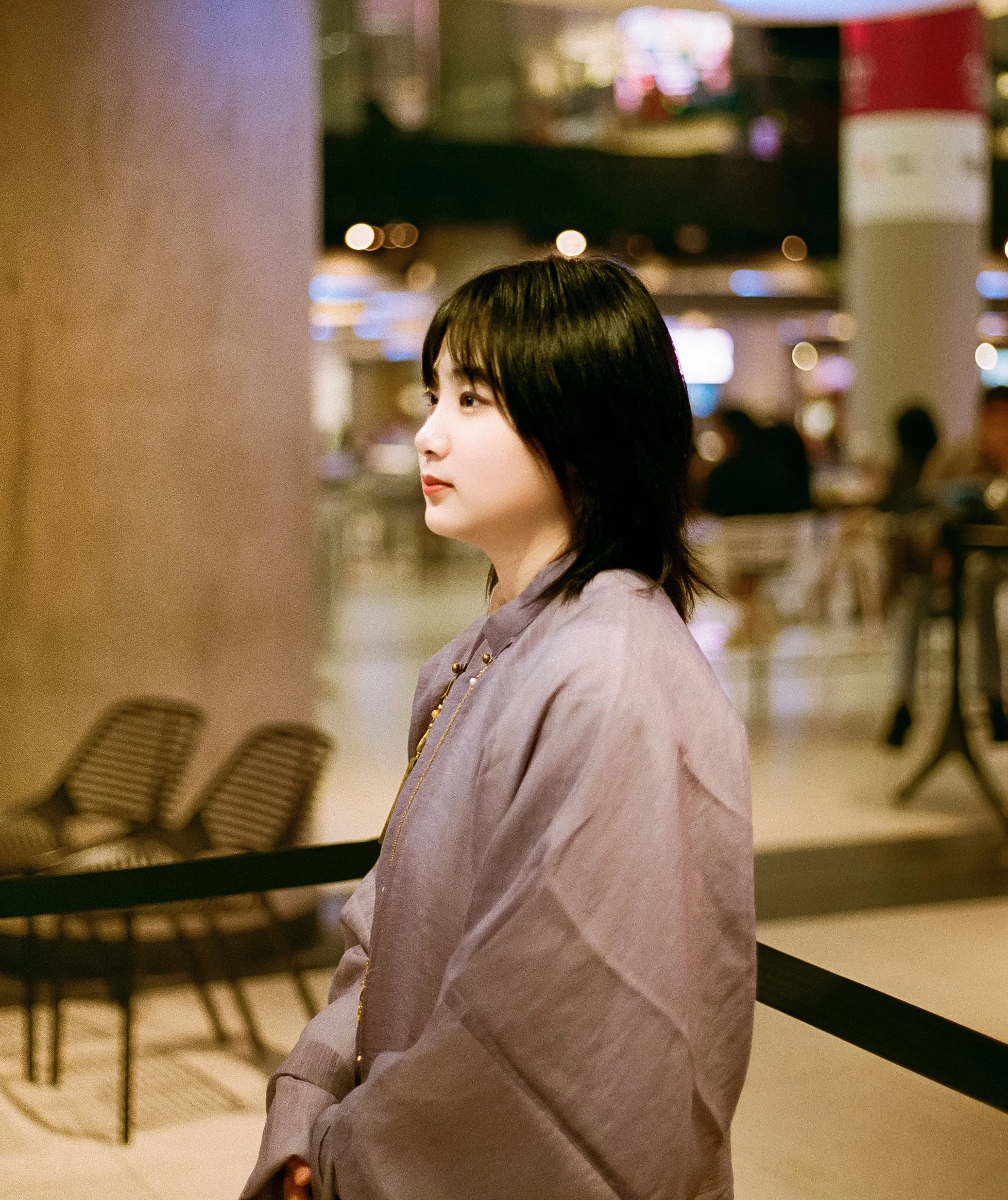 A young woman with shoulder-length black hair stands in an indoor public space, possibly an airport or shopping mall, looking to the left. She is wearing a light-colored, oversized jacket and a necklace, with a blurry background of chairs, people, an