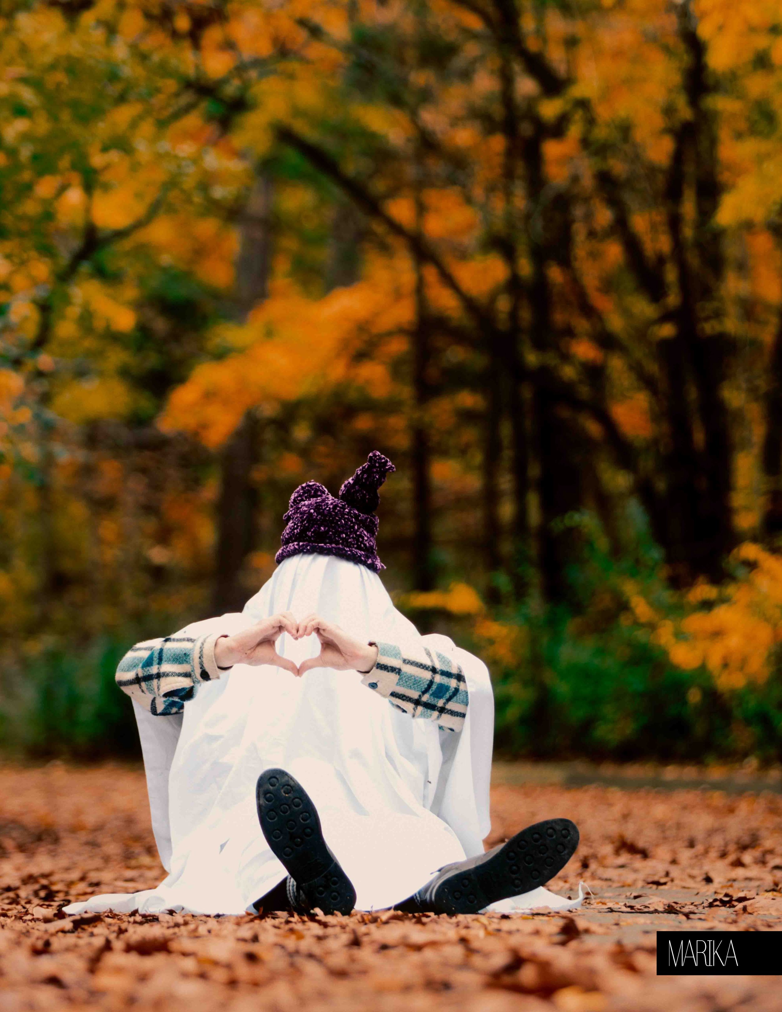 Person sitting outdoors on fallen autumn leaves, making a heart shape with their hands, wearing a purple knit hat with a top knot, a patterned coat, and a white sheet or blanket covering their head, in front of a forest with orange and yellow foliage