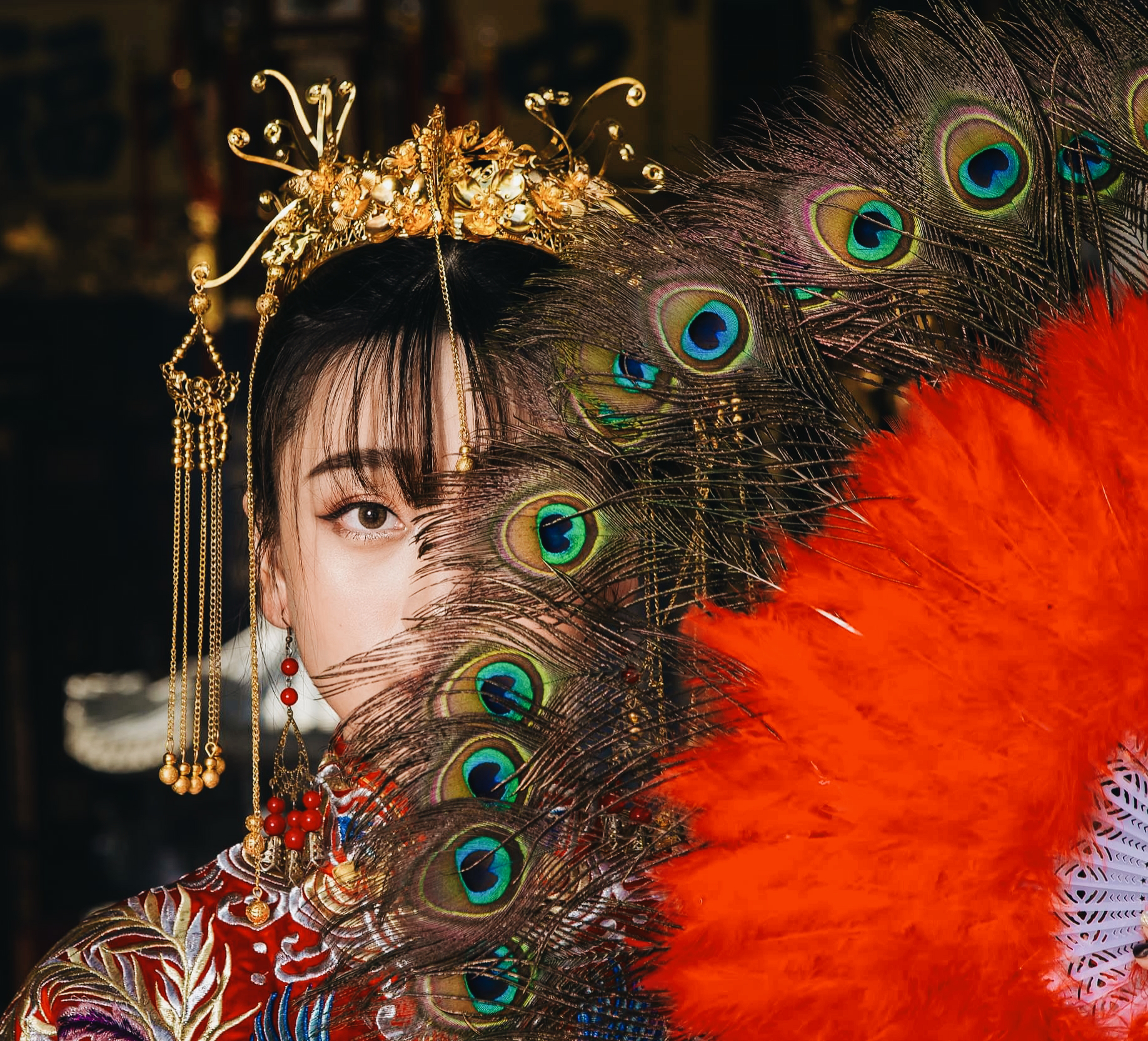 A woman dressed in traditional Asian attire, holding a fan with peacock feathers and orange feathers, wearing an ornate gold headpiece and jewelry.
