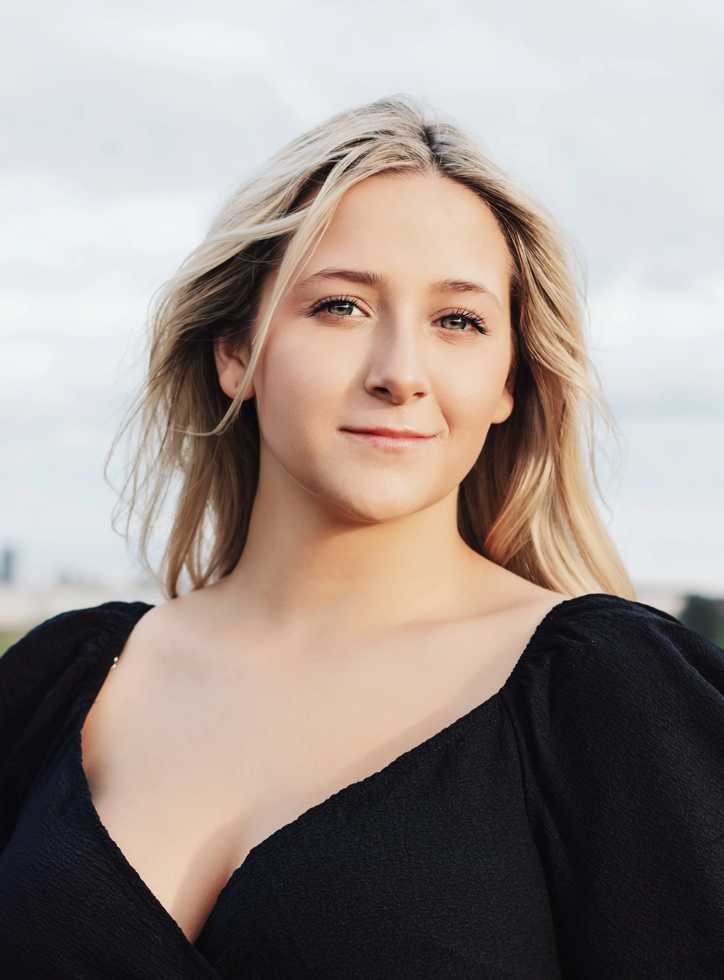 A portrait of a young woman with blonde hair, light skin, wearing a black top, smiling slightly, with a cloudy sky in the background.