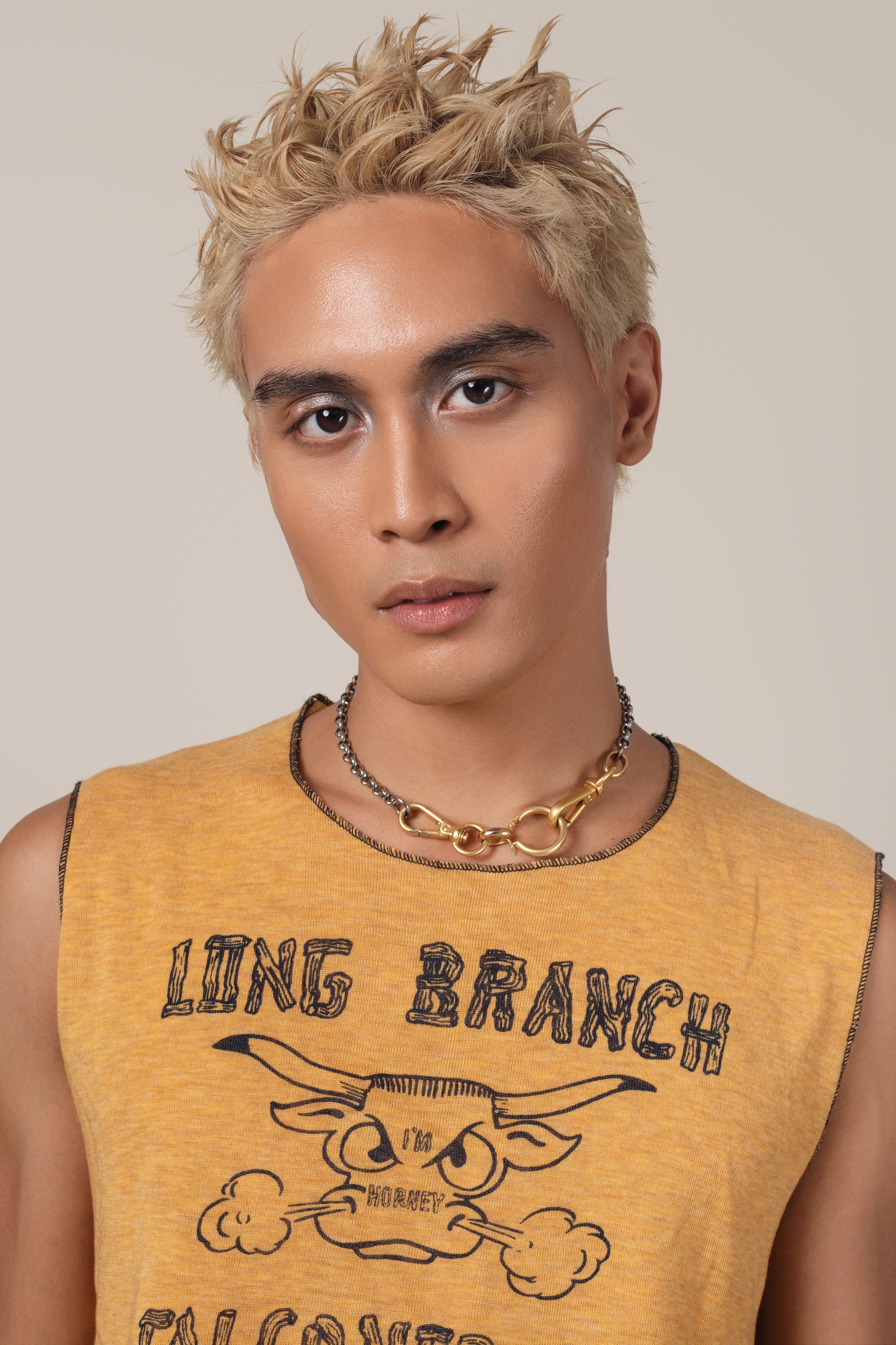 A young person with platinum blonde, spiky hair wearing a sleeveless yellow T-shirt with a graphic of a horned animal and the text "Long Branch" and "Honey." They have a silver and gold chain necklace and a neutral expression, looking directly at the
