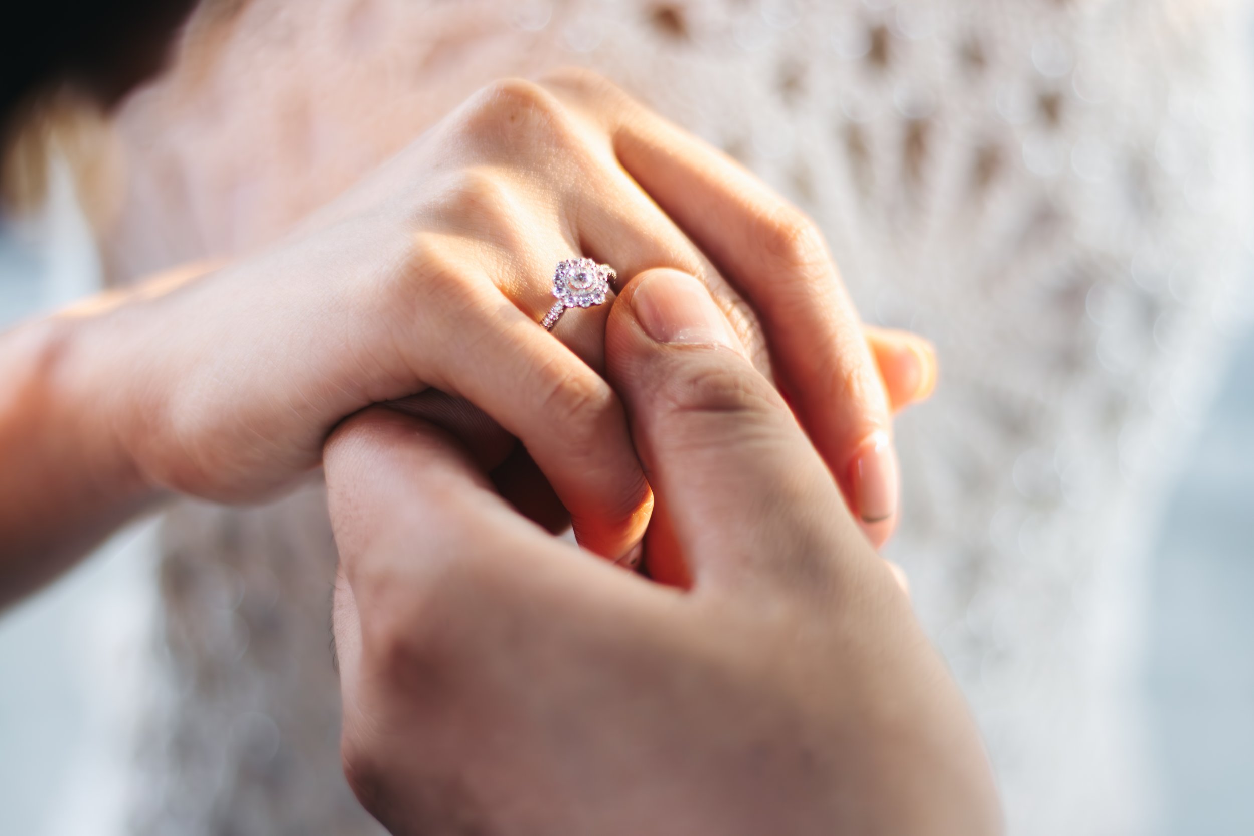 Close-up of a person’s hand showing a wedding or engagement ring with a large central stone, held by another hand against a blurred background.