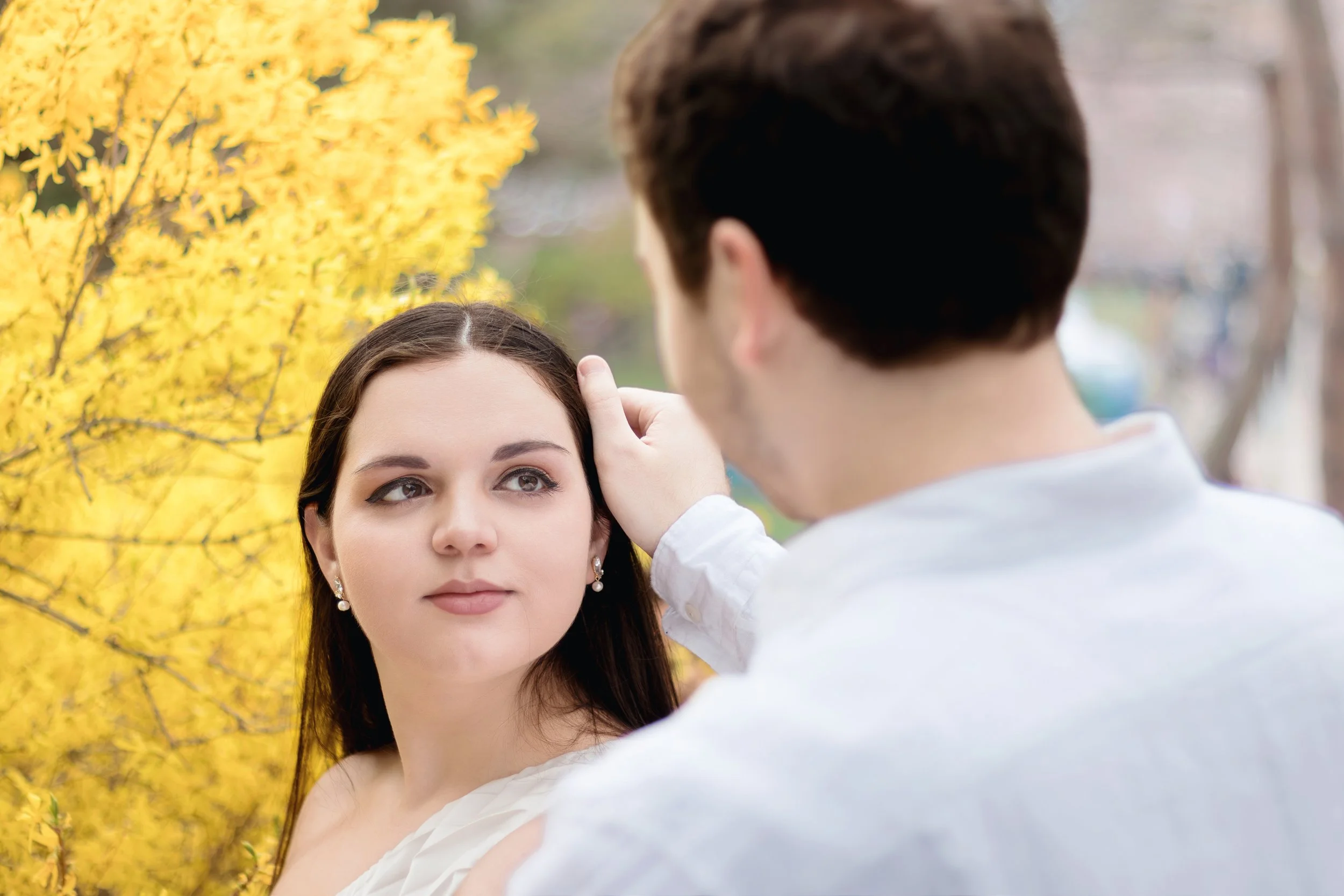 A man gently touching a woman's forehead outdoors with yellow flowering bushes in the background during daytime.