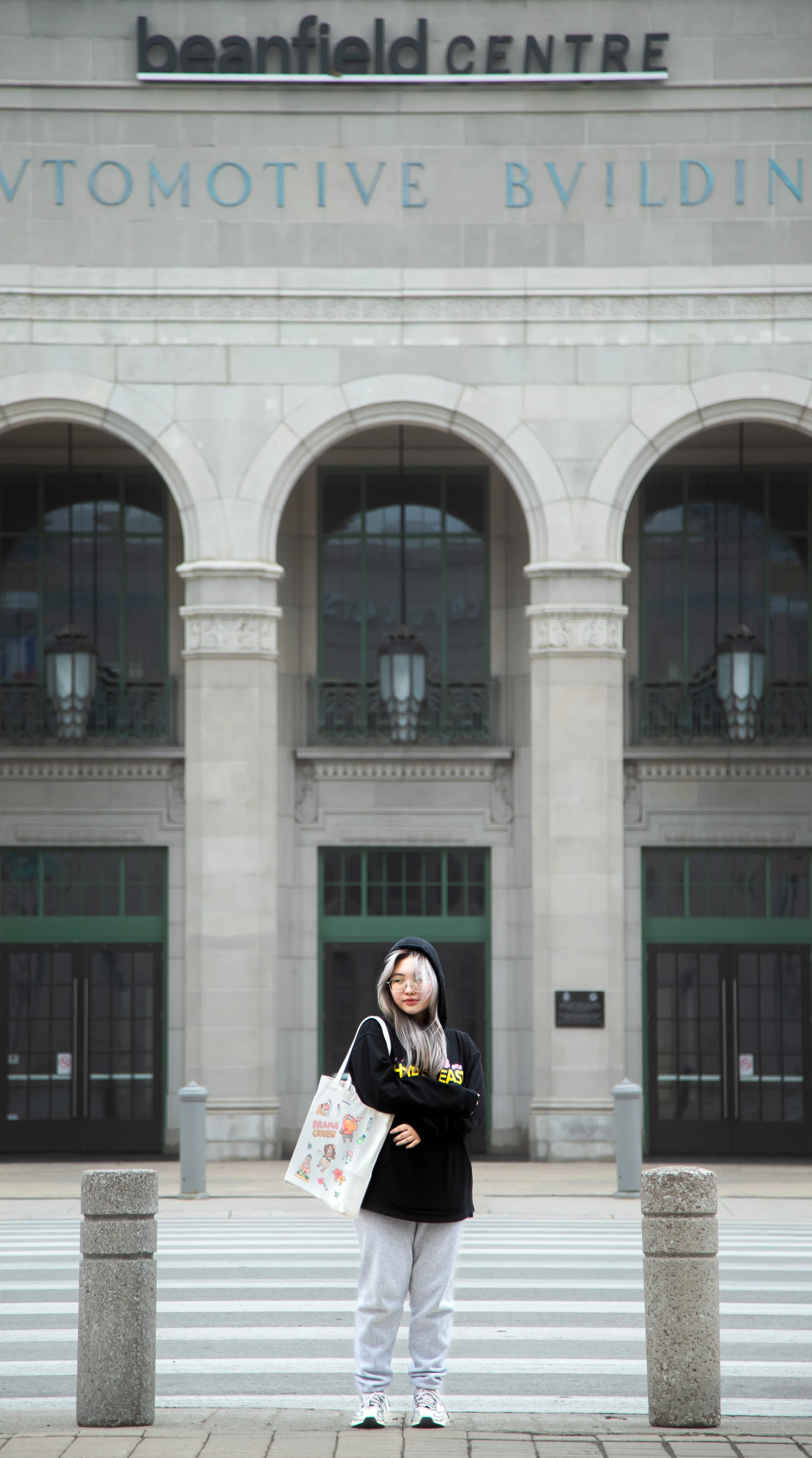 A young woman with gray hair, glasses, and a black hoodie standing on a crosswalk in front of the Beanfield Centre, a large historic building with arched windows and decorative stonework, holding a tote bag with colorful illustrations.