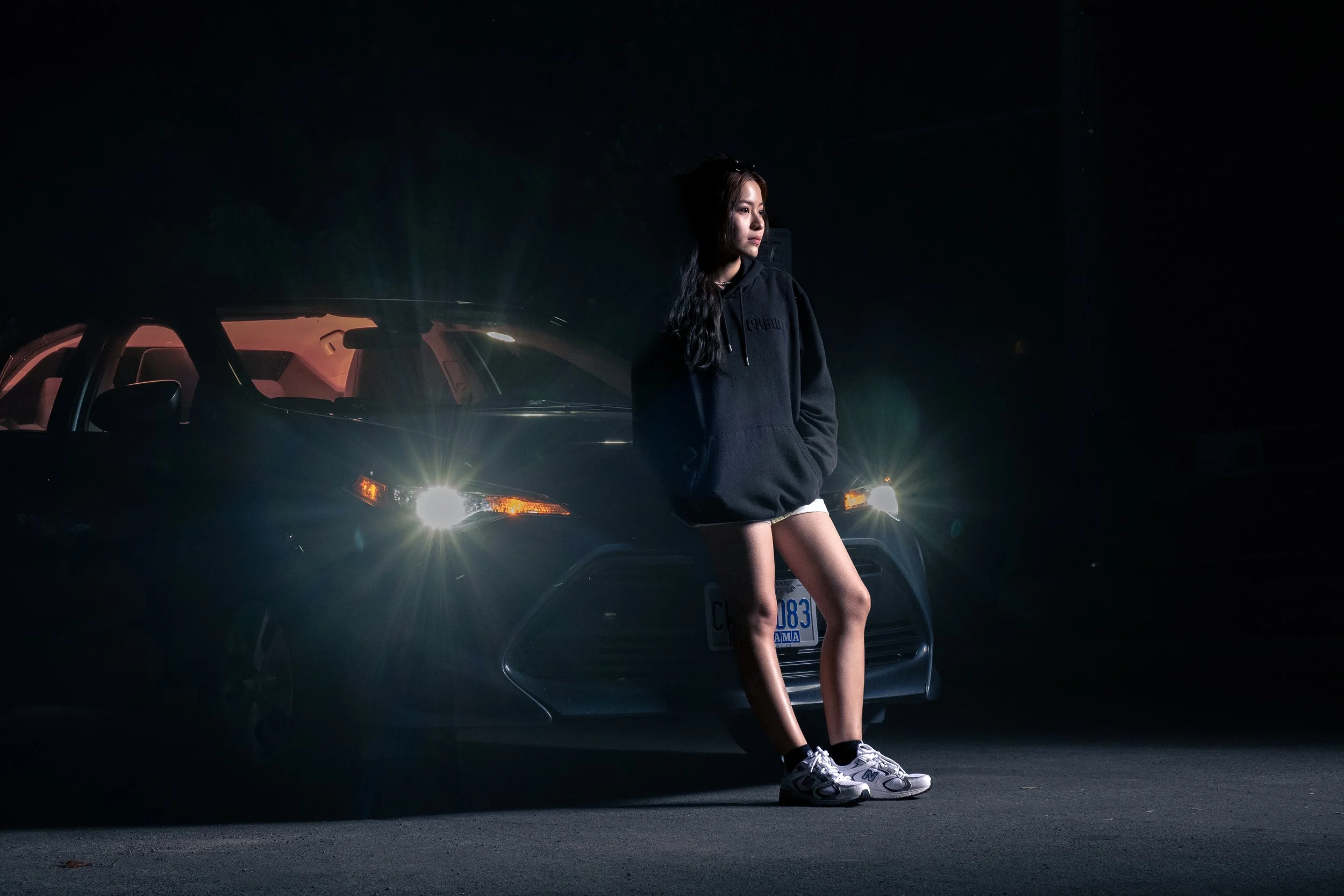 A young woman standing in front of a dark car at night, with the headlights on and casting light.