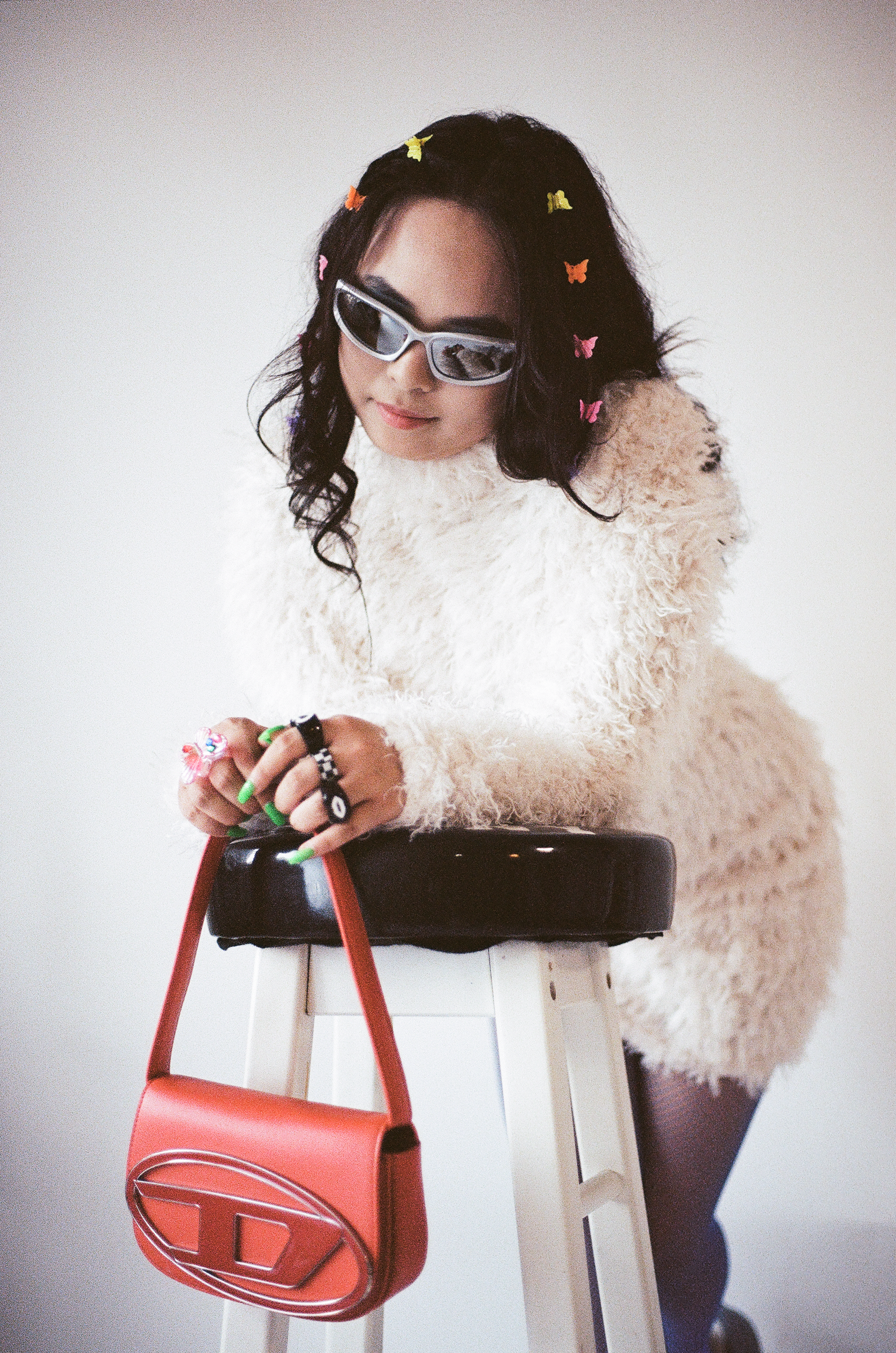 A woman with dark wavy hair adorned with small colorful butterfly clips, wearing reflective sunglasses and a fluffy cream-colored sweater, leaning on a stool with a small orange handbag hanging from it.