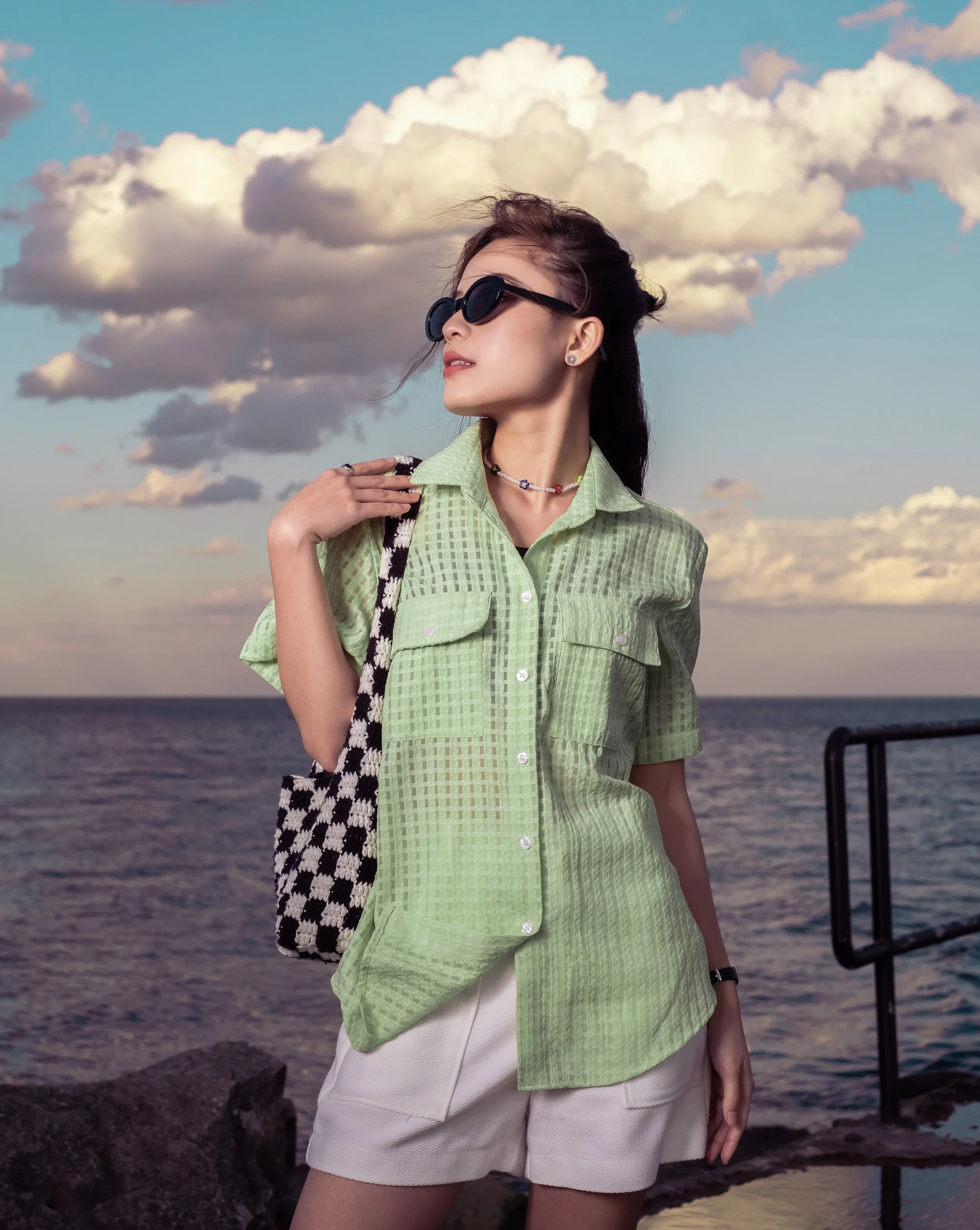 Woman wearing black sunglasses and a green checkered shirt with pockets, holding a black and white checkered bag, standing on rocks near the ocean with cloud-filled sky in the background.