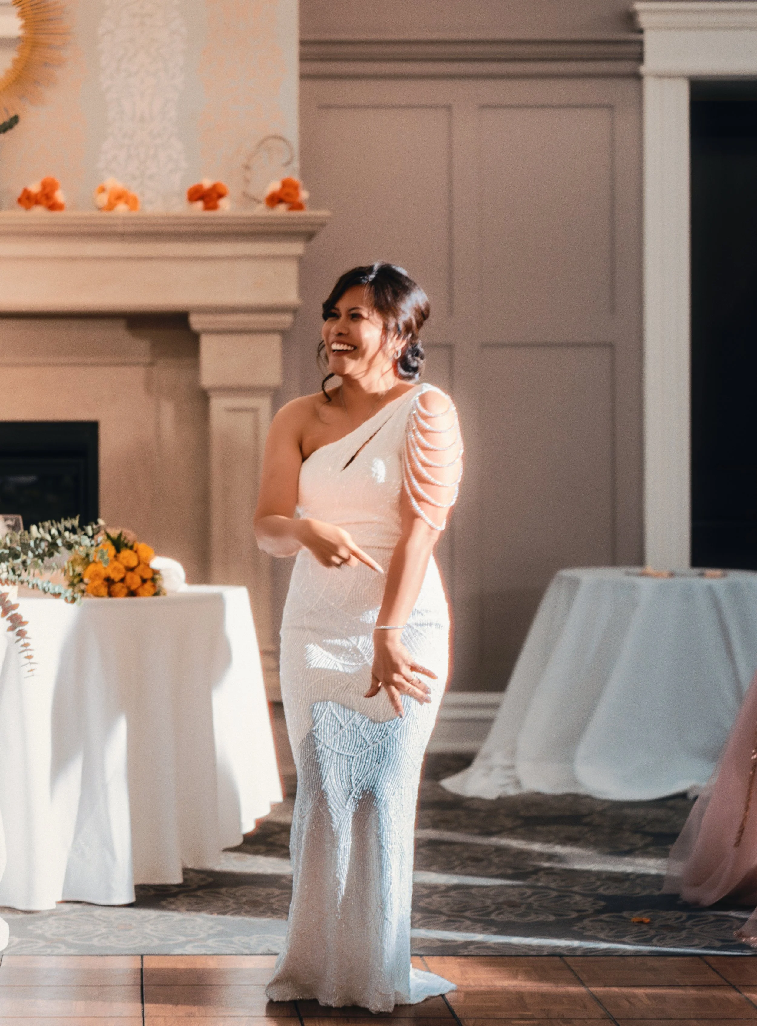 A woman in a one-shoulder white dress with decorative pearl shoulder details, smiling and pointing her finger, standing in a decorated room with tables and flowers.