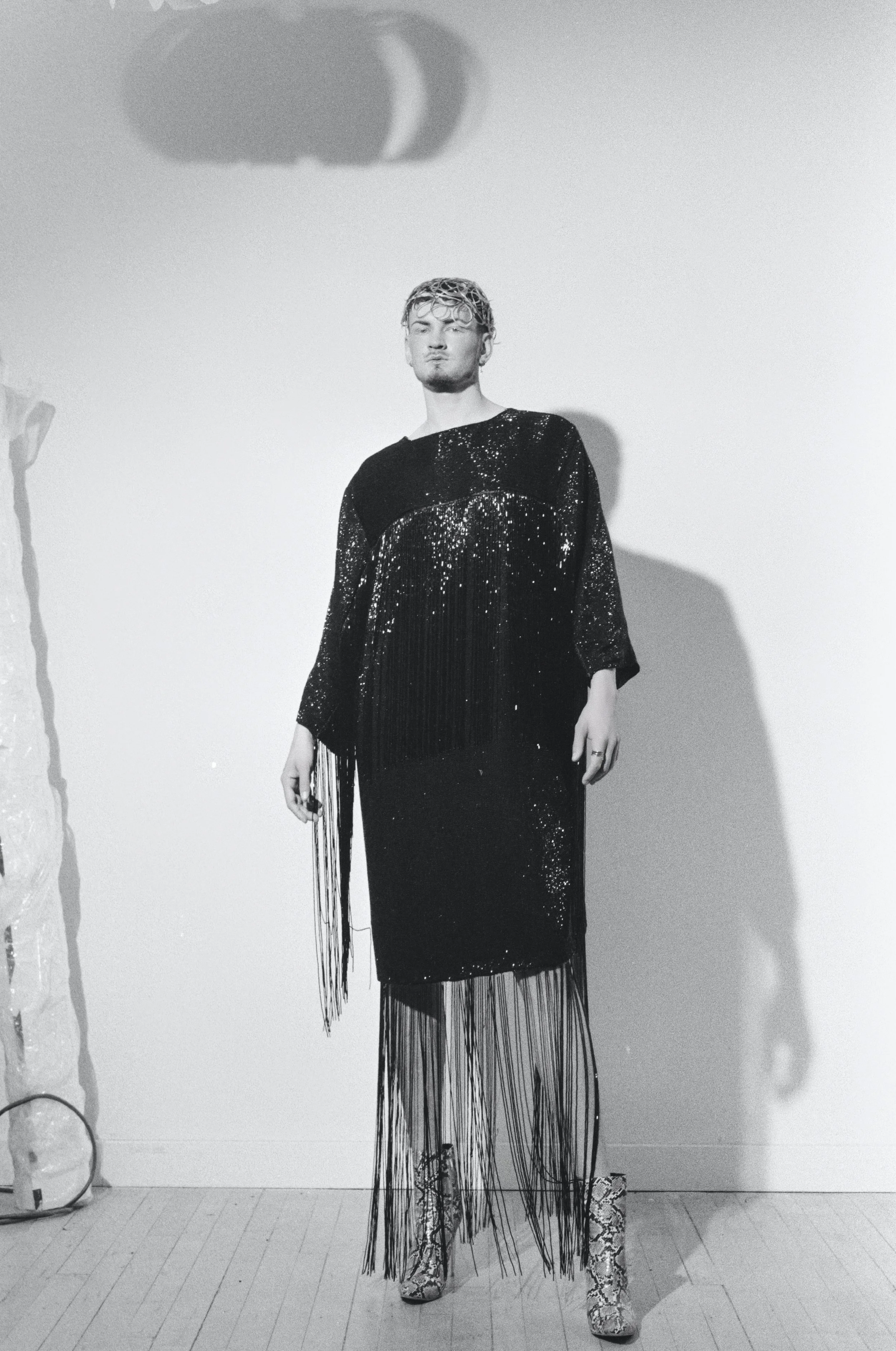 A model wearing a black sequined dress with long fringe details, standing on display.