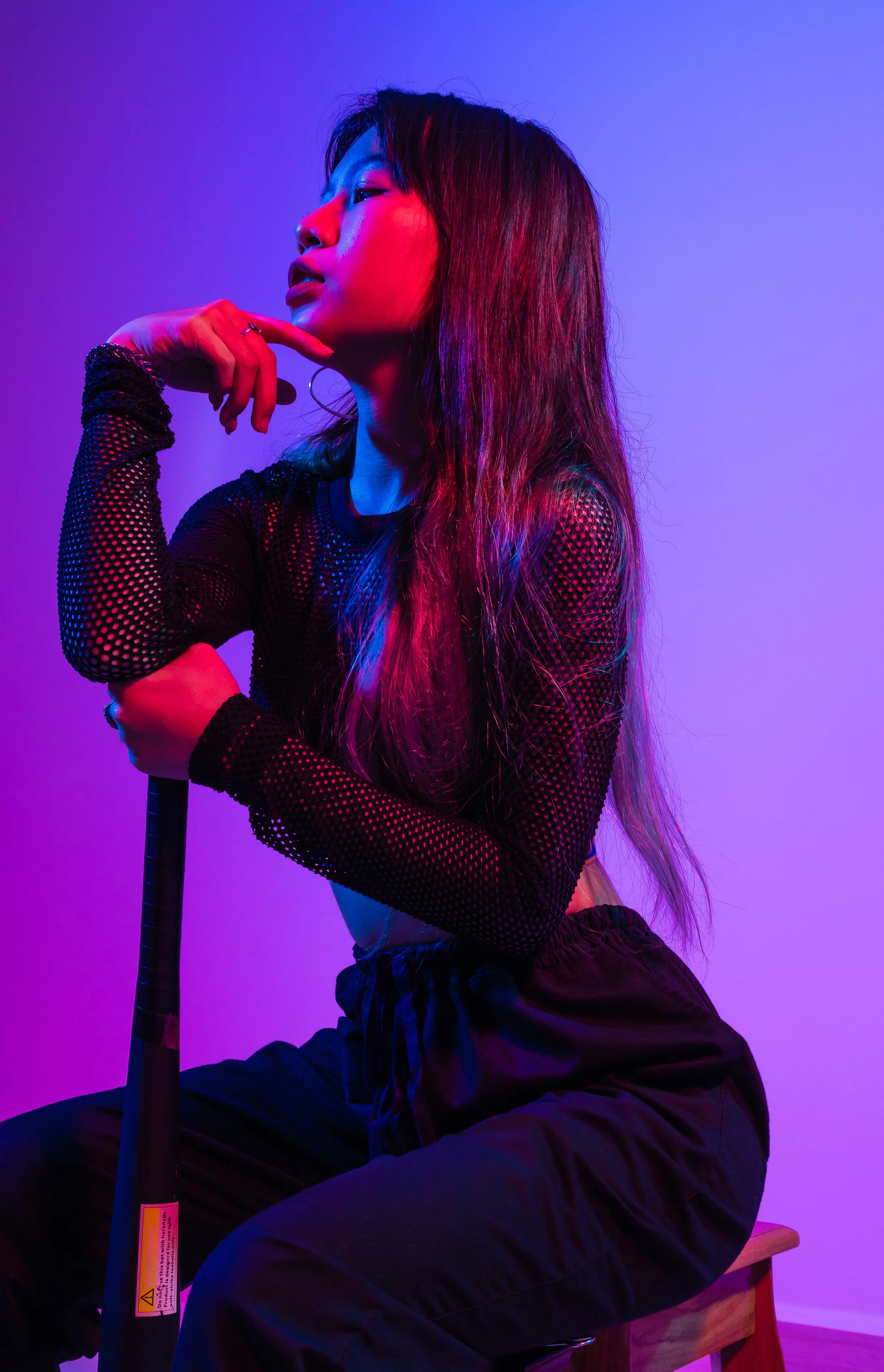 A young woman with long hair sits on a stool, dressed in a black mesh top and black pants, illuminated by vibrant purple and pink lighting.