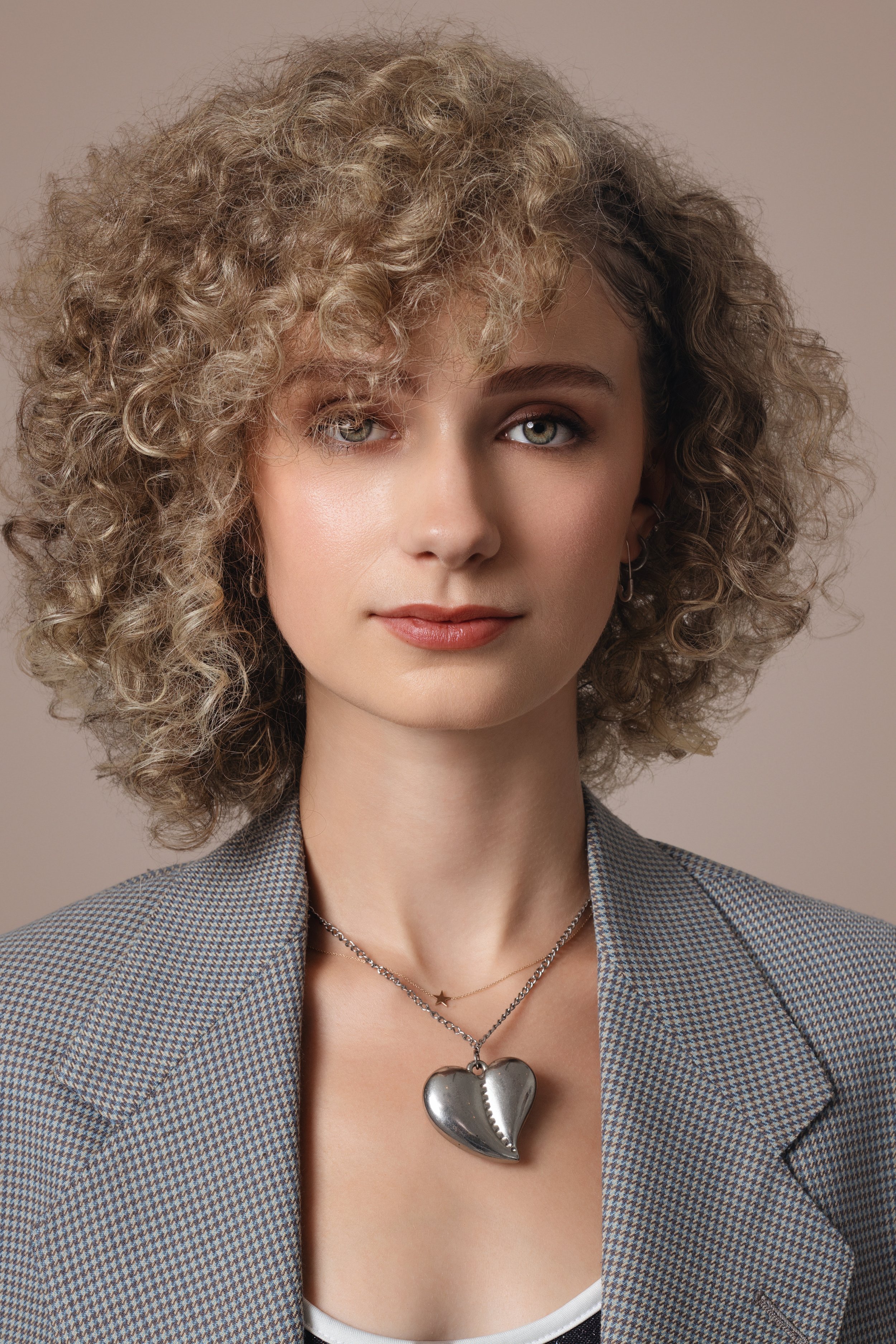 A woman with blonde curly hair wearing a gray checkered blazer and a silver heart-shaped necklace.