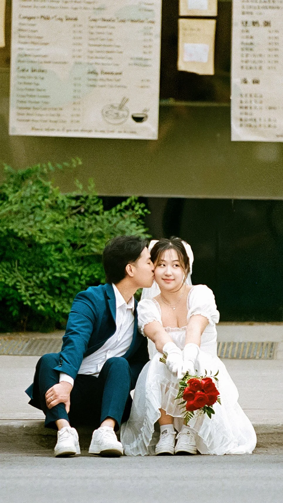A couple dressed in wedding attire, with the groom in a blue suit and the bride in a white dress, sitting on the pavement. The groom is kissing the bride on the cheek, and she is holding a bouquet of red roses while smiling and looking at the camera.