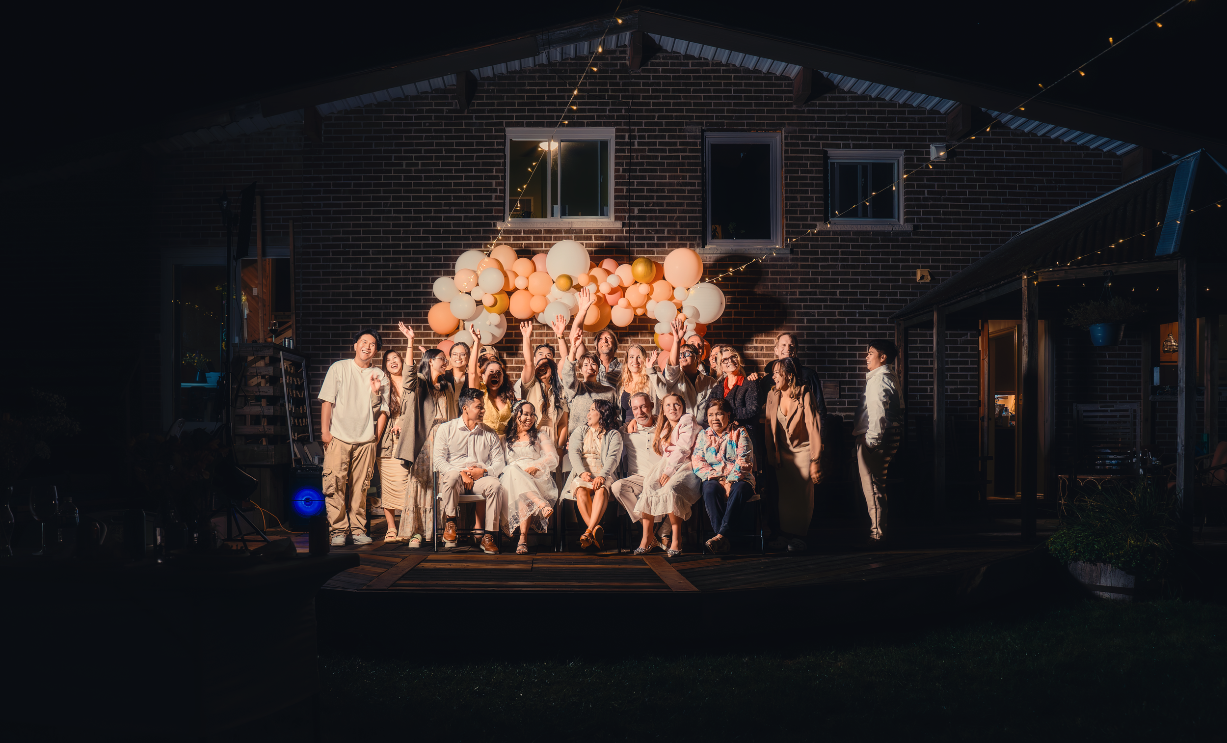 Group of people celebrating outdoors at night with balloons and string lights, posing for a photo in front of a brick house.
