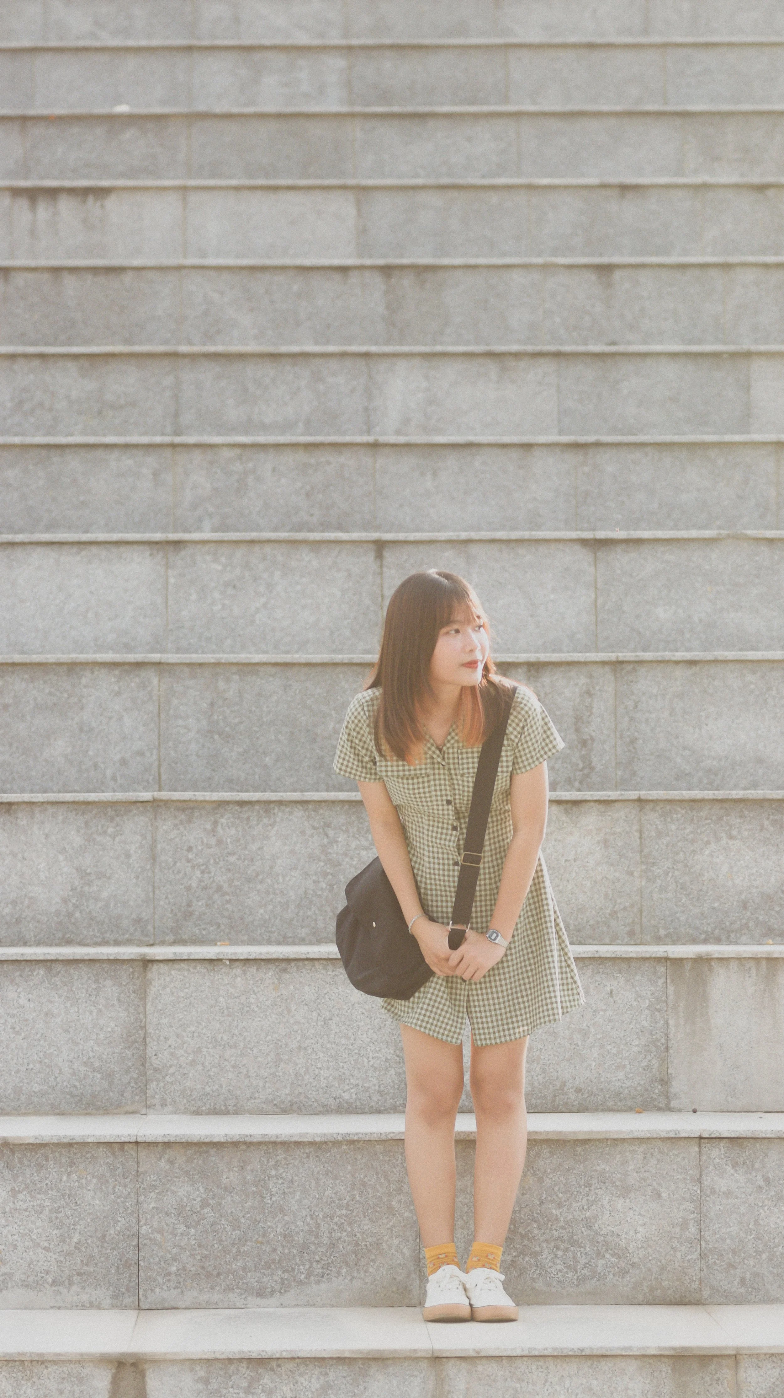 A young woman with shoulder-length hair, wearing a green checkered dress, white sneakers, yellow socks, and a black shoulder bag, stands on outdoor steps made of gray stone, looking to the side.