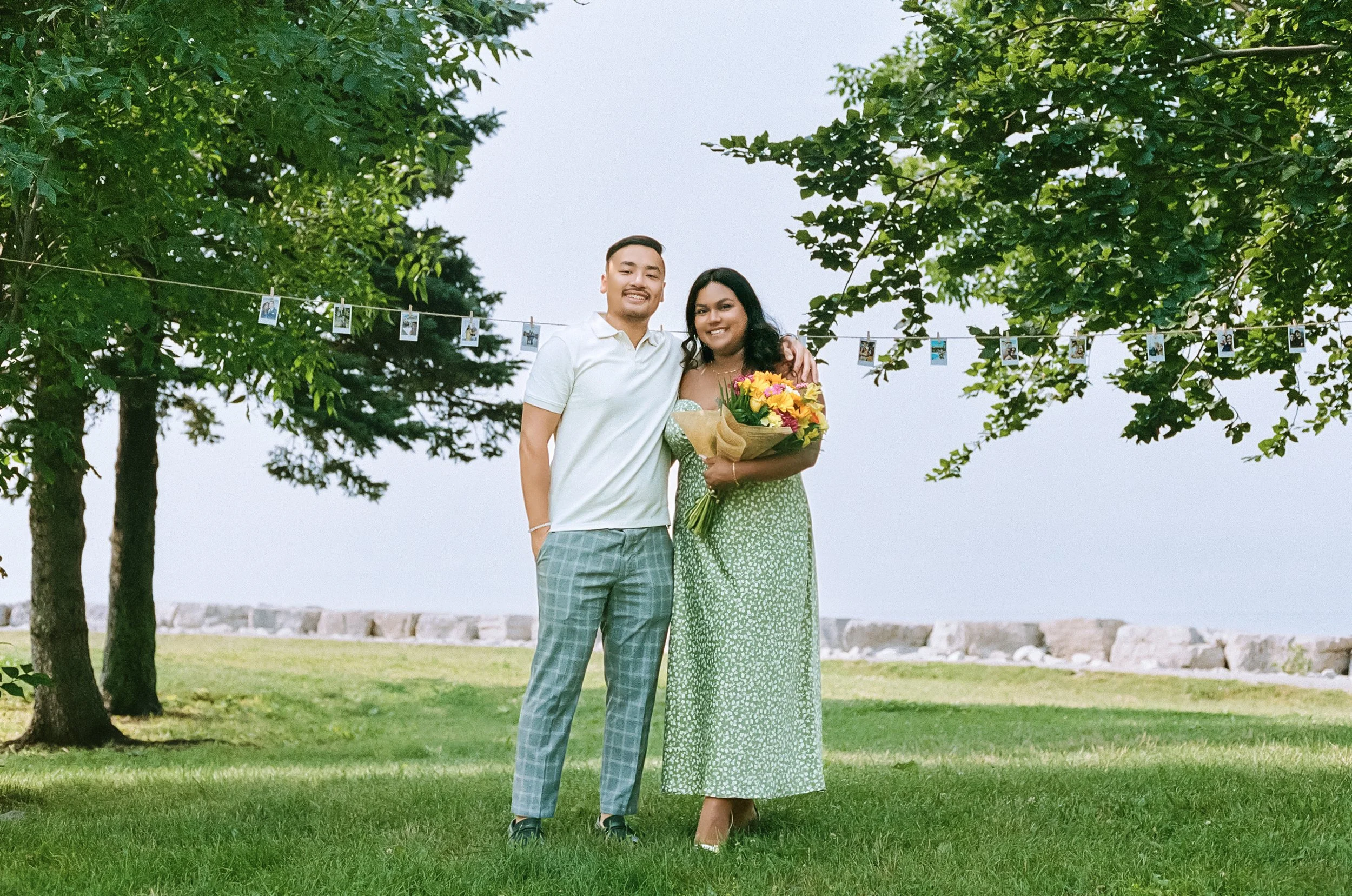 A couple standing on a grassy area with trees and a body of water in the background, the woman holding a bouquet of flowers.