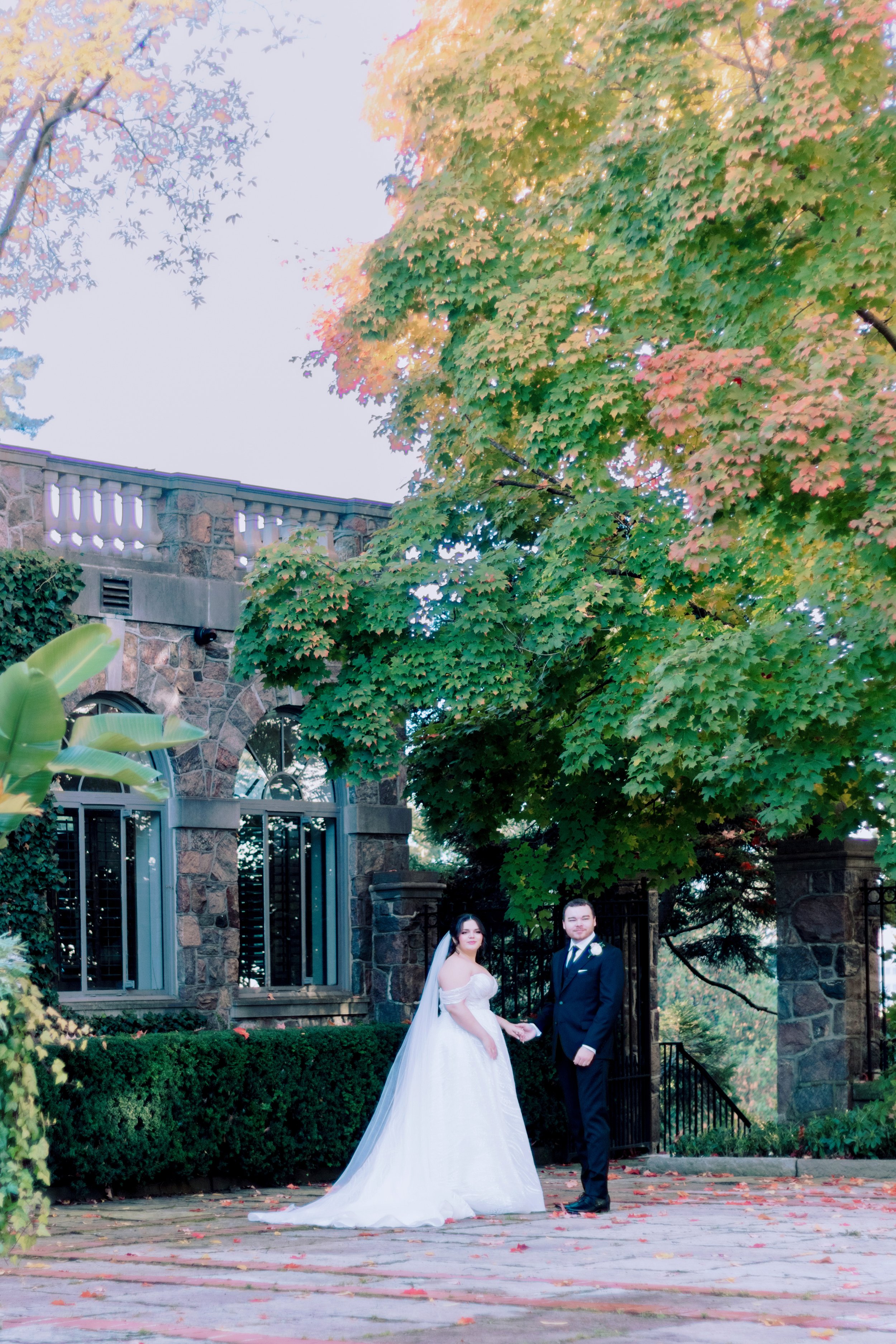 A bride and groom holding hands outside under a large tree with colorful fall foliage at a wedding.