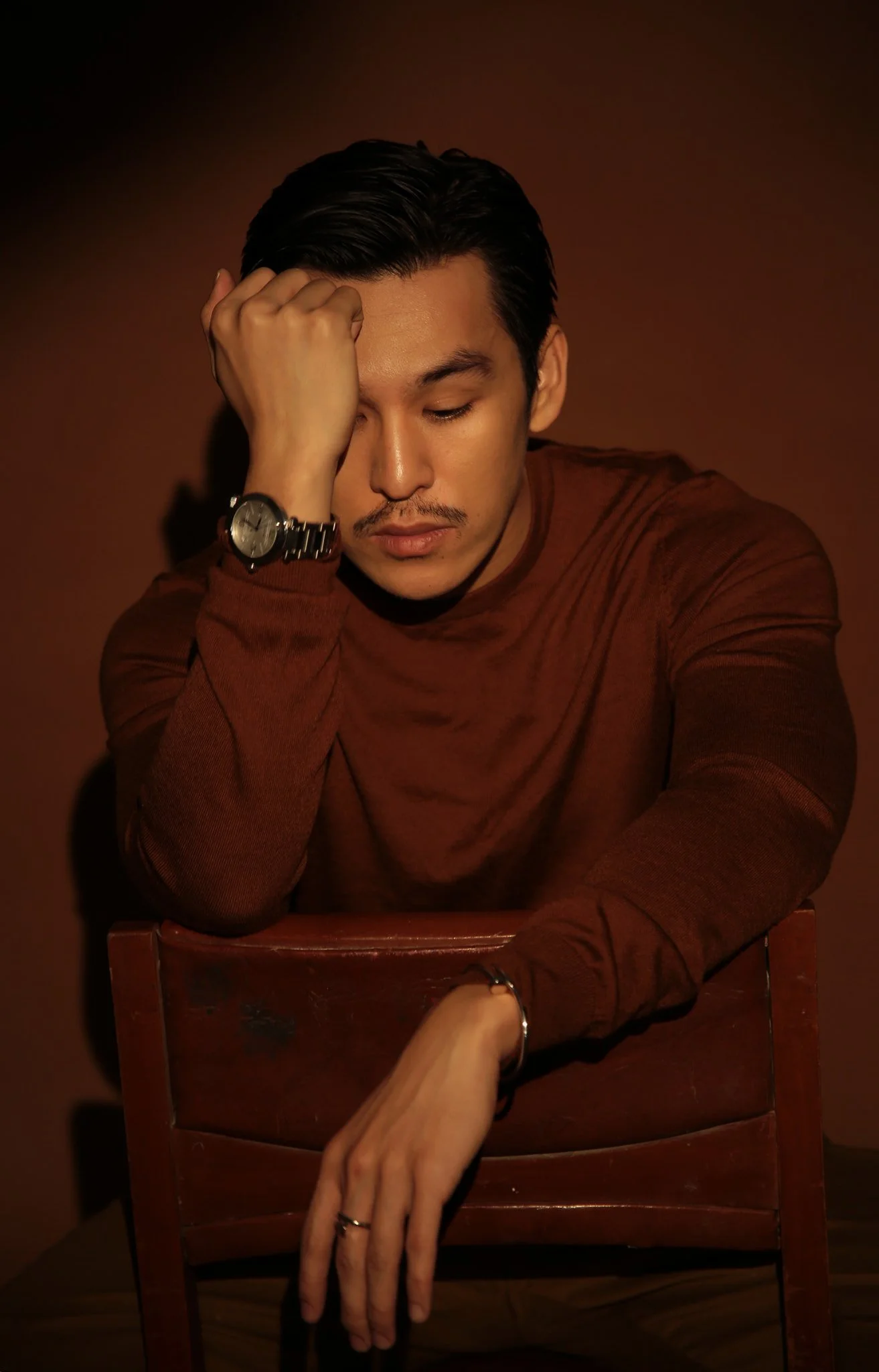 A man with dark hair and a mustache, wearing a brown long-sleeve shirt, sitting with his eyes closed, resting his forehead on his hand, with a wristwatch on his left wrist, sitting behind a wooden chair, against a brown background.
