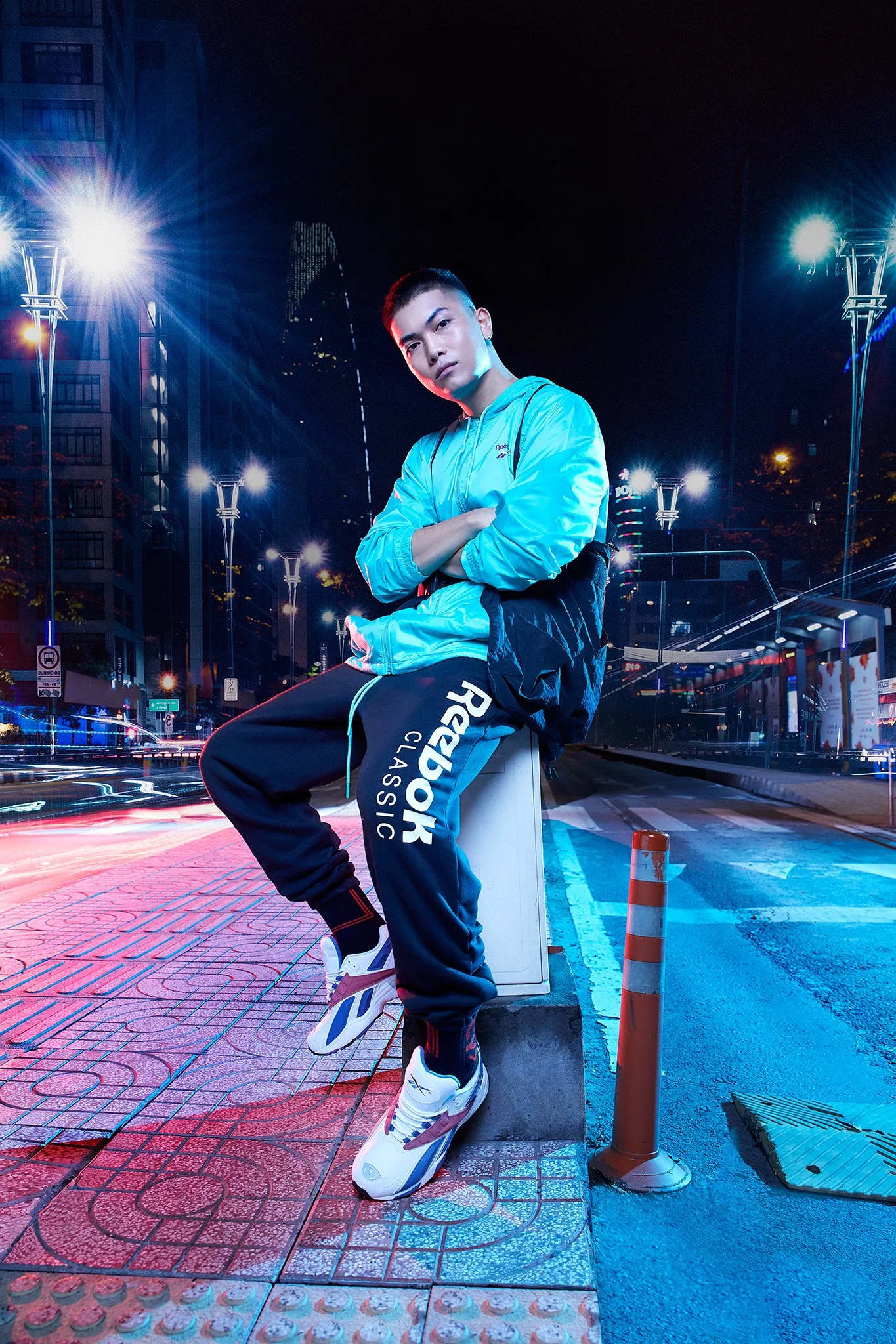 Young man in athletic clothing sitting on a city street at night, with bright streetlights and buildings in the background.