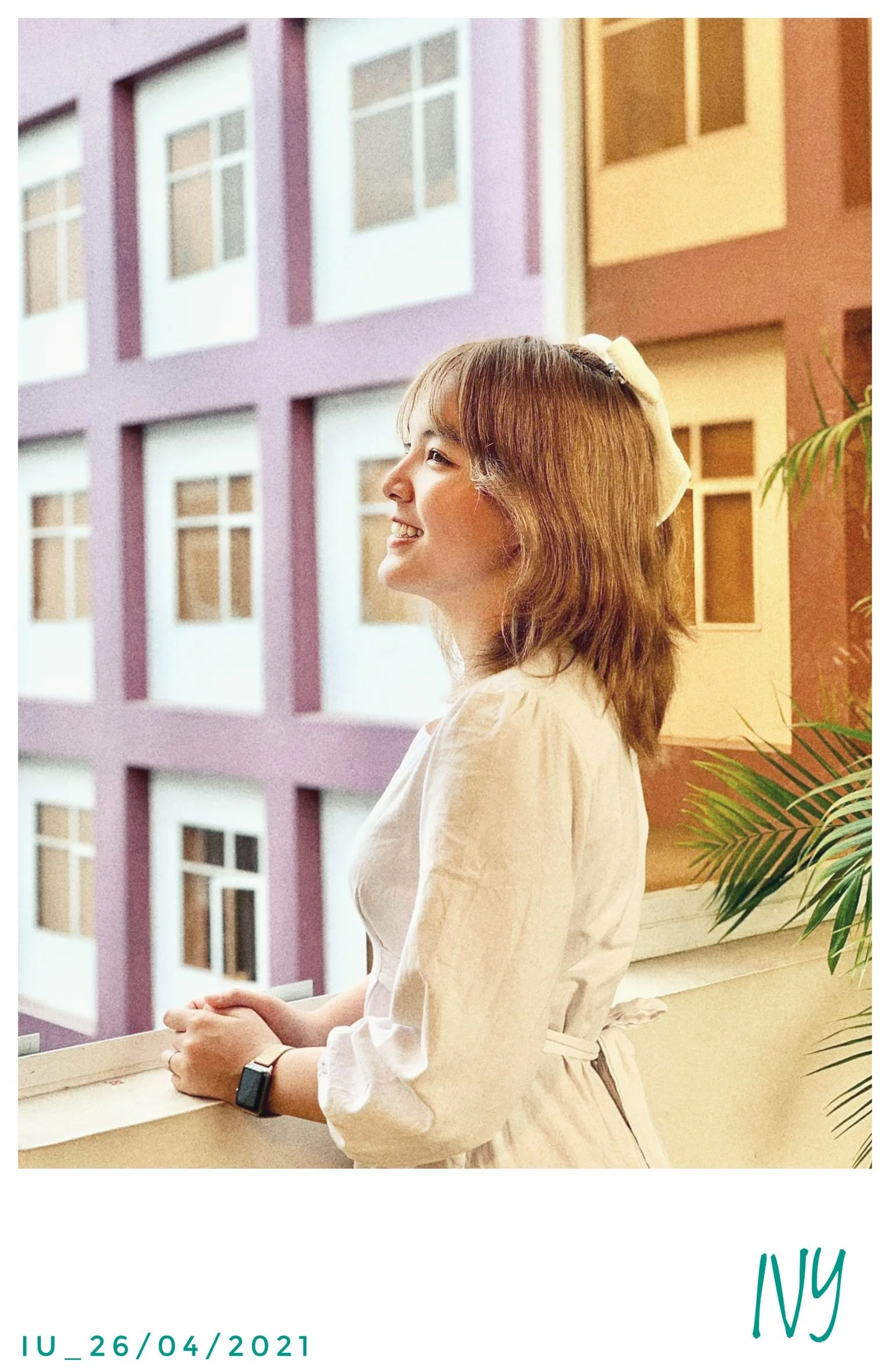 A young woman with shoulder-length brown hair, wearing a white dress and a watch, standing on a balcony with a smile, looking to her left. Behind her is a colorful apartment building with purple, yellow, and beige sections and multiple windows.