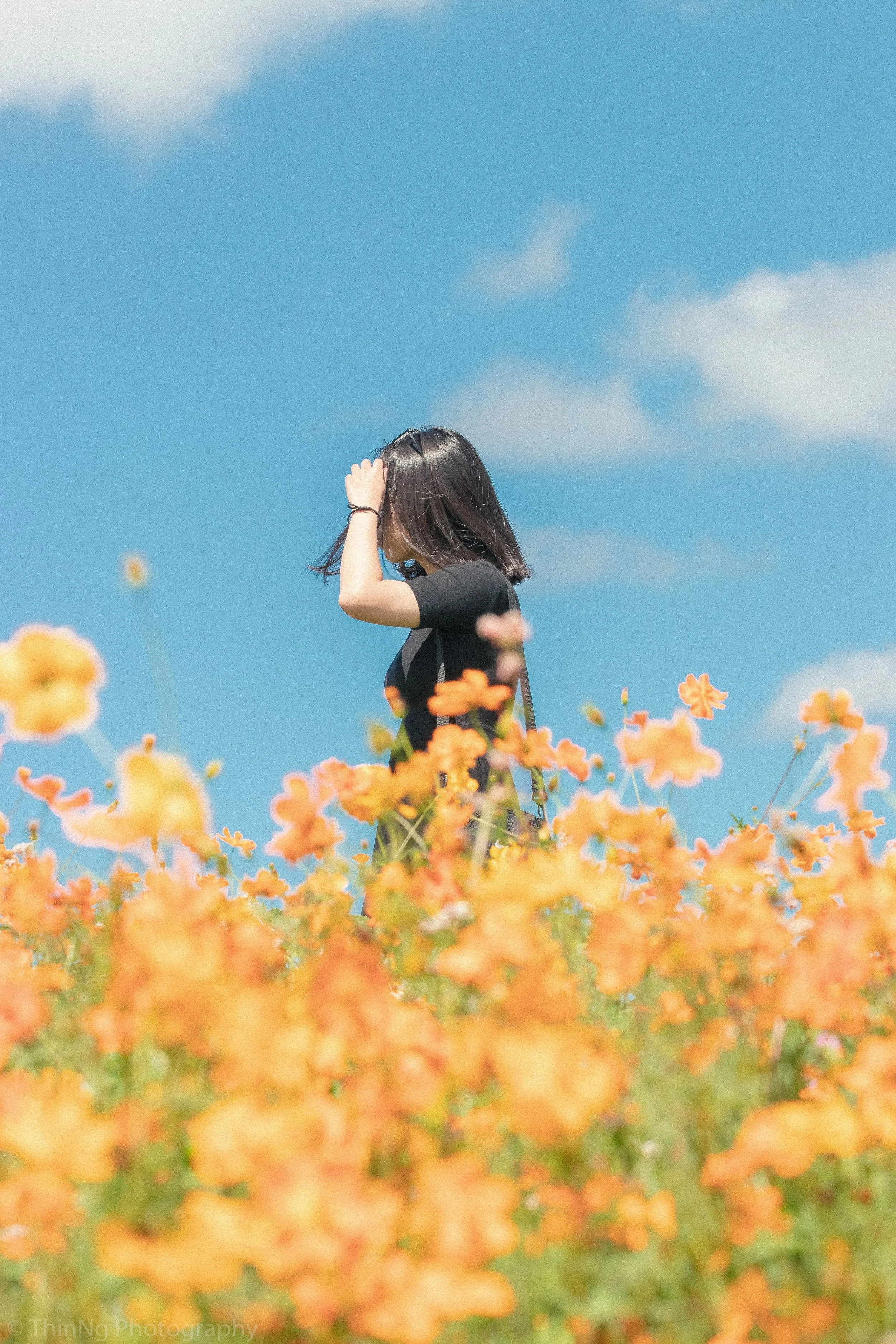 A woman with black hair wearing a black T-shirt standing in a field of orange flowers under a blue sky with scattered clouds.