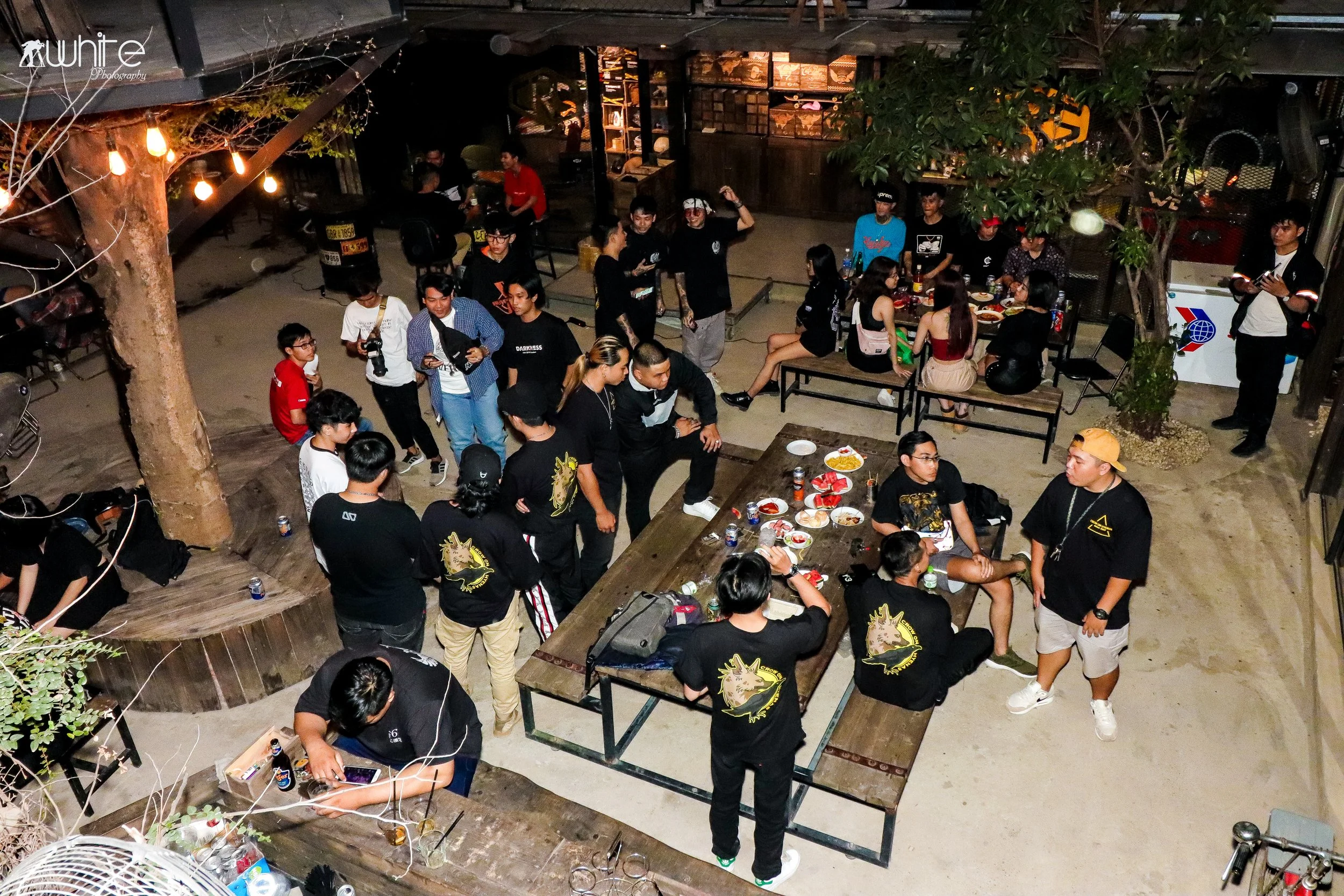 A group of people gathered at an outdoor night event, standing, sitting, and talking around tables with food and drinks, with some engaging with their phones and others socializing.