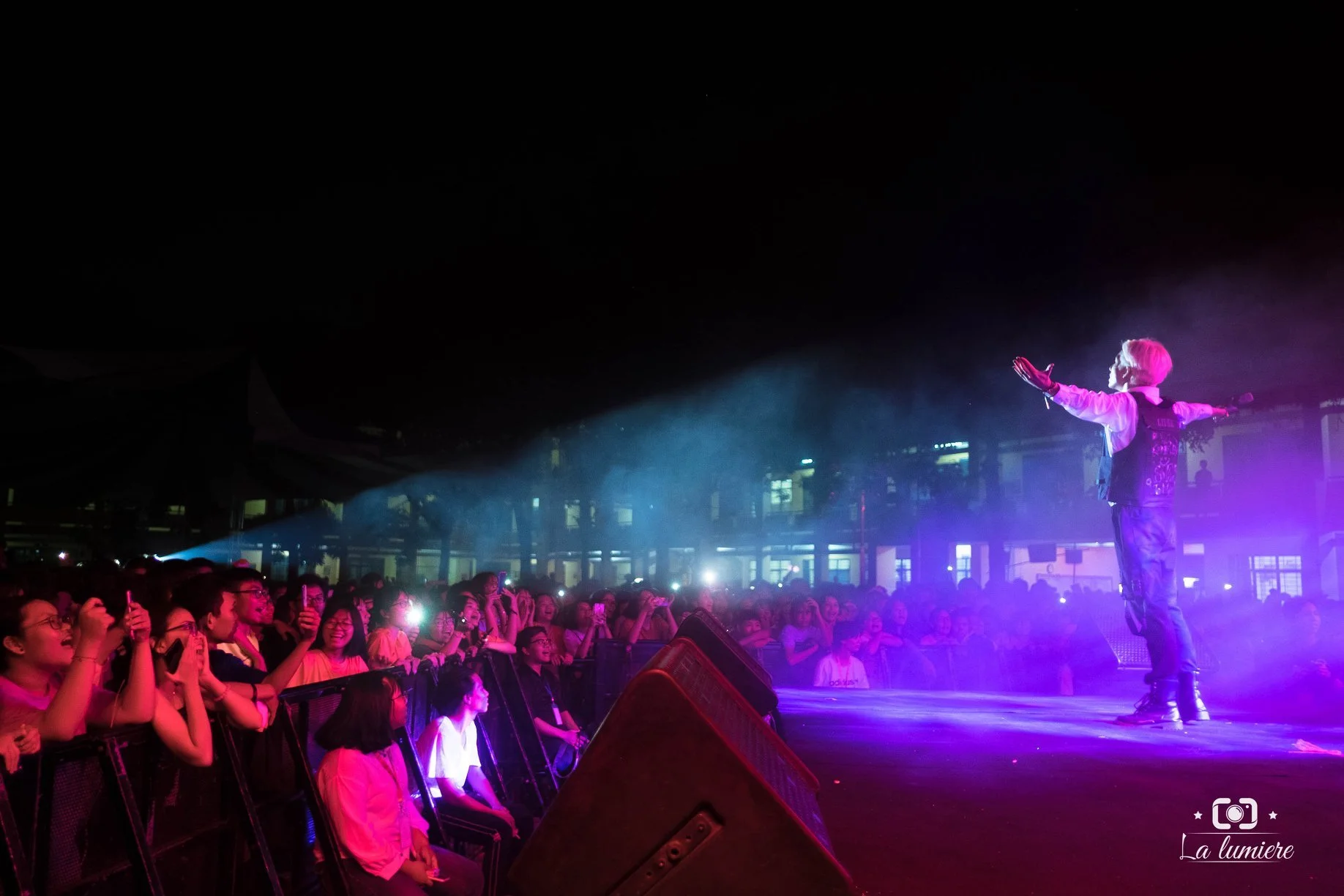 Performer with pink hair on stage with arms outstretched singing at a concert, with a large crowd in front illuminated by stage lights.