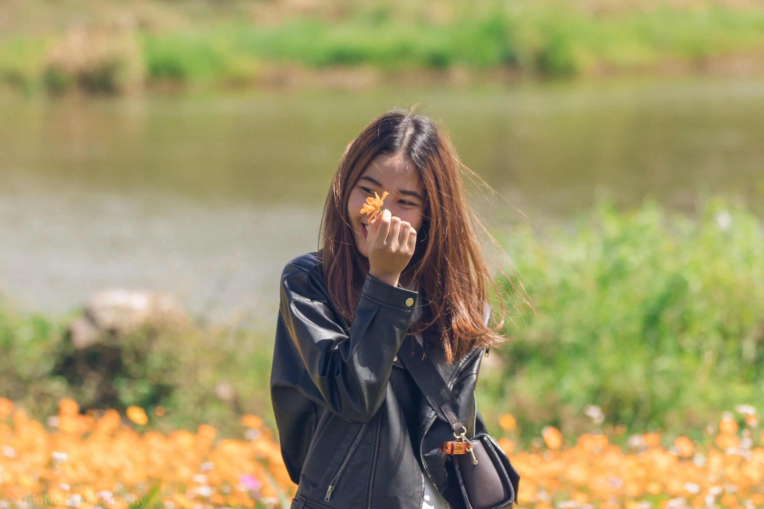 A woman smiling and holding a small orange flower near her nose outdoors with a lake and green grassy area in the background.