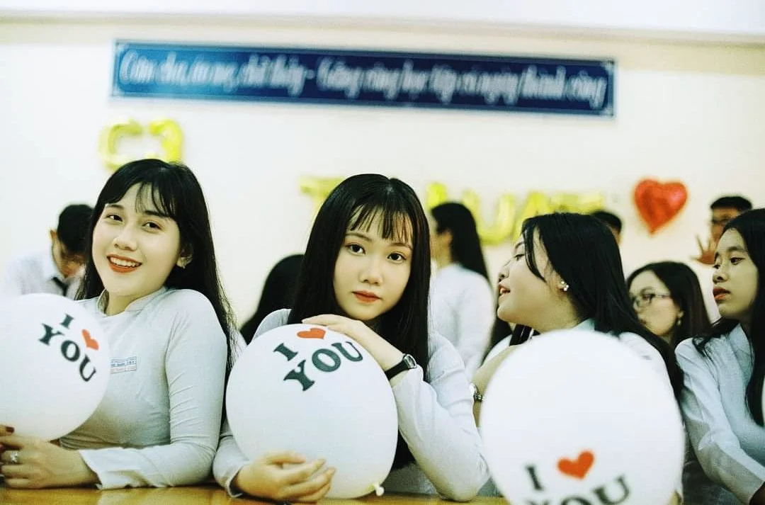 Group of young women sitting at a table with white balloons that say "I love you." They are in a decorated room with balloons and a banner, likely at a celebration or event.