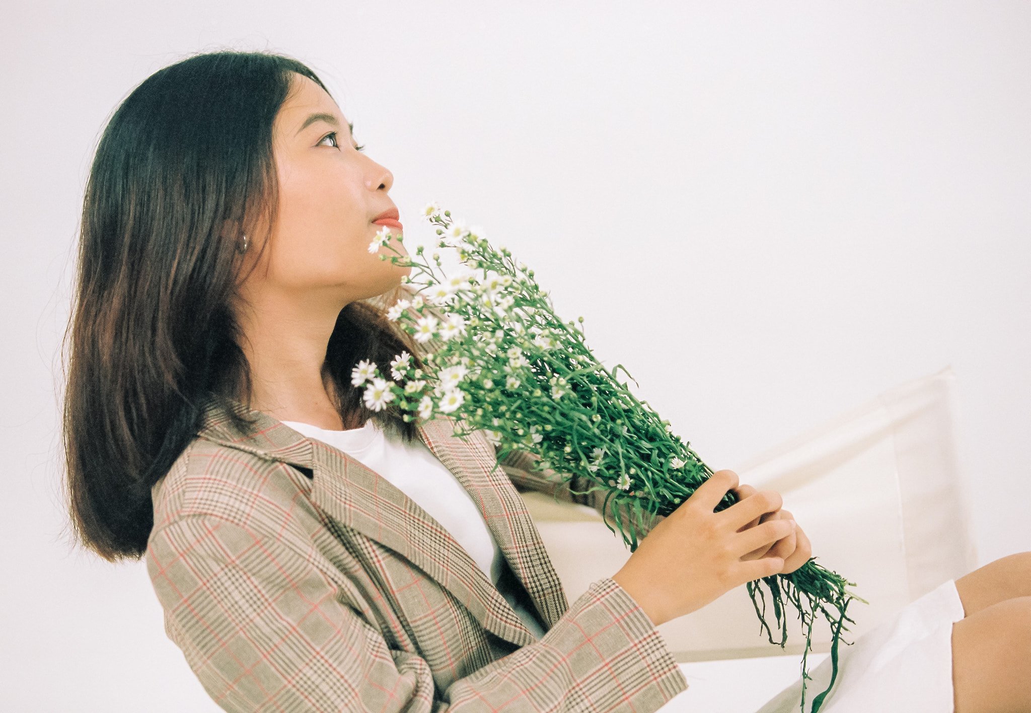 A woman with dark hair wearing a plaid blazer and white top is holding a large bouquet of small white flowers with green stems and leaves.