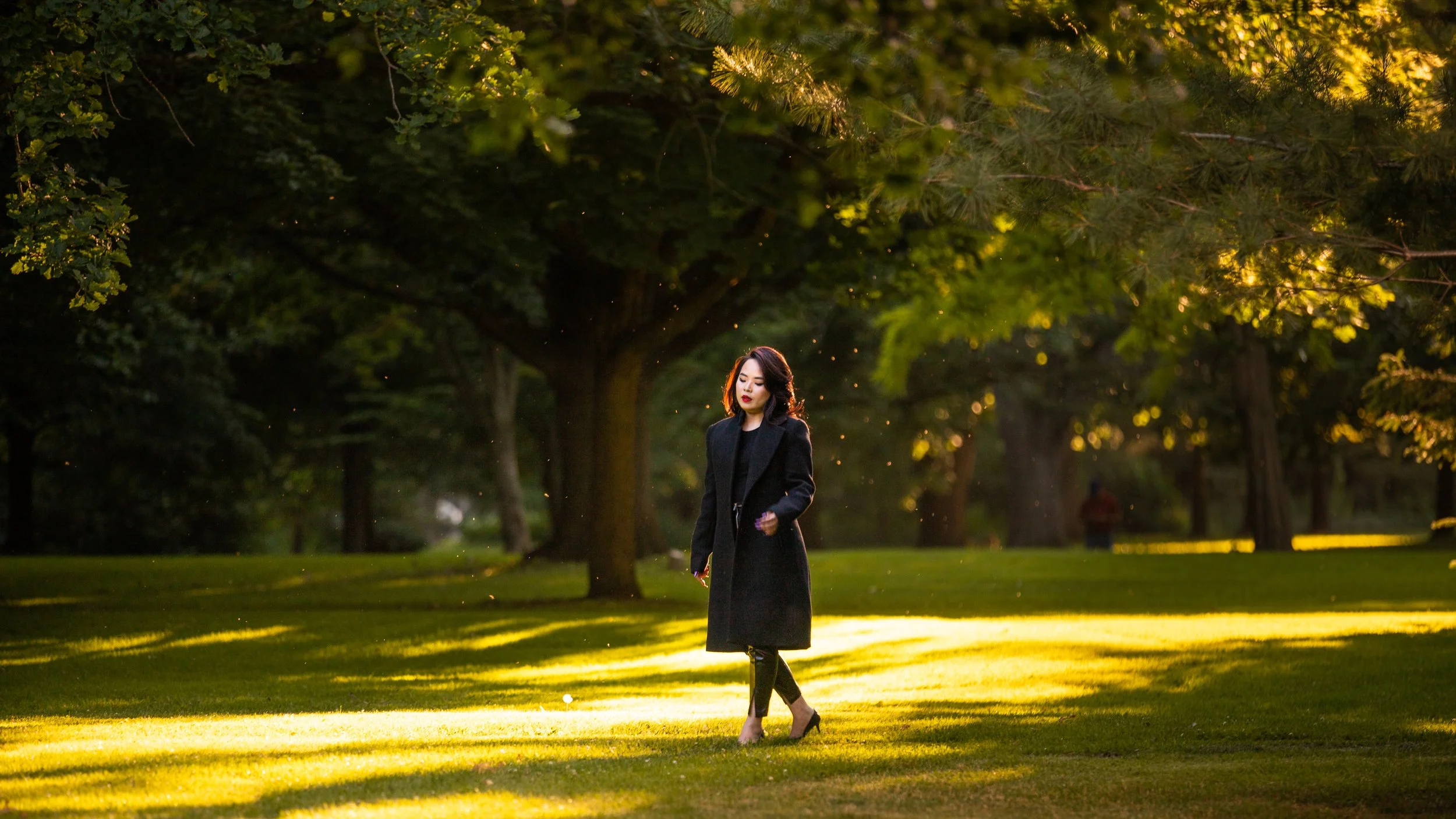 A woman walking in a park at sunset, dressed in a black coat and black pants, with trees surrounding her and sunlight filtering through the branches.