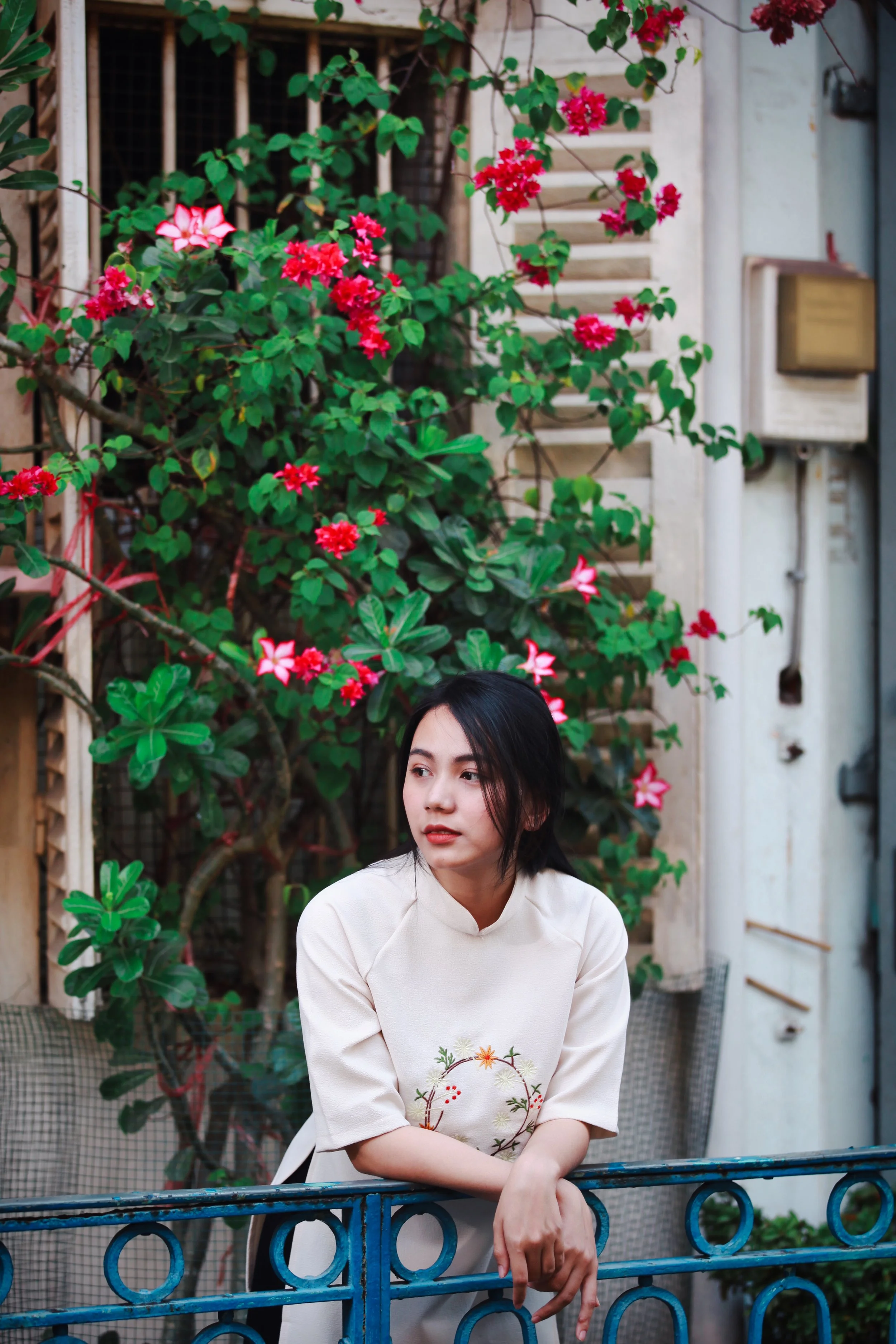 A young woman in a cream-colored dress with floral embroidery resting her arms on a blue railing, standing in front of vibrant pink and red flower bushes and greenery outdoors.