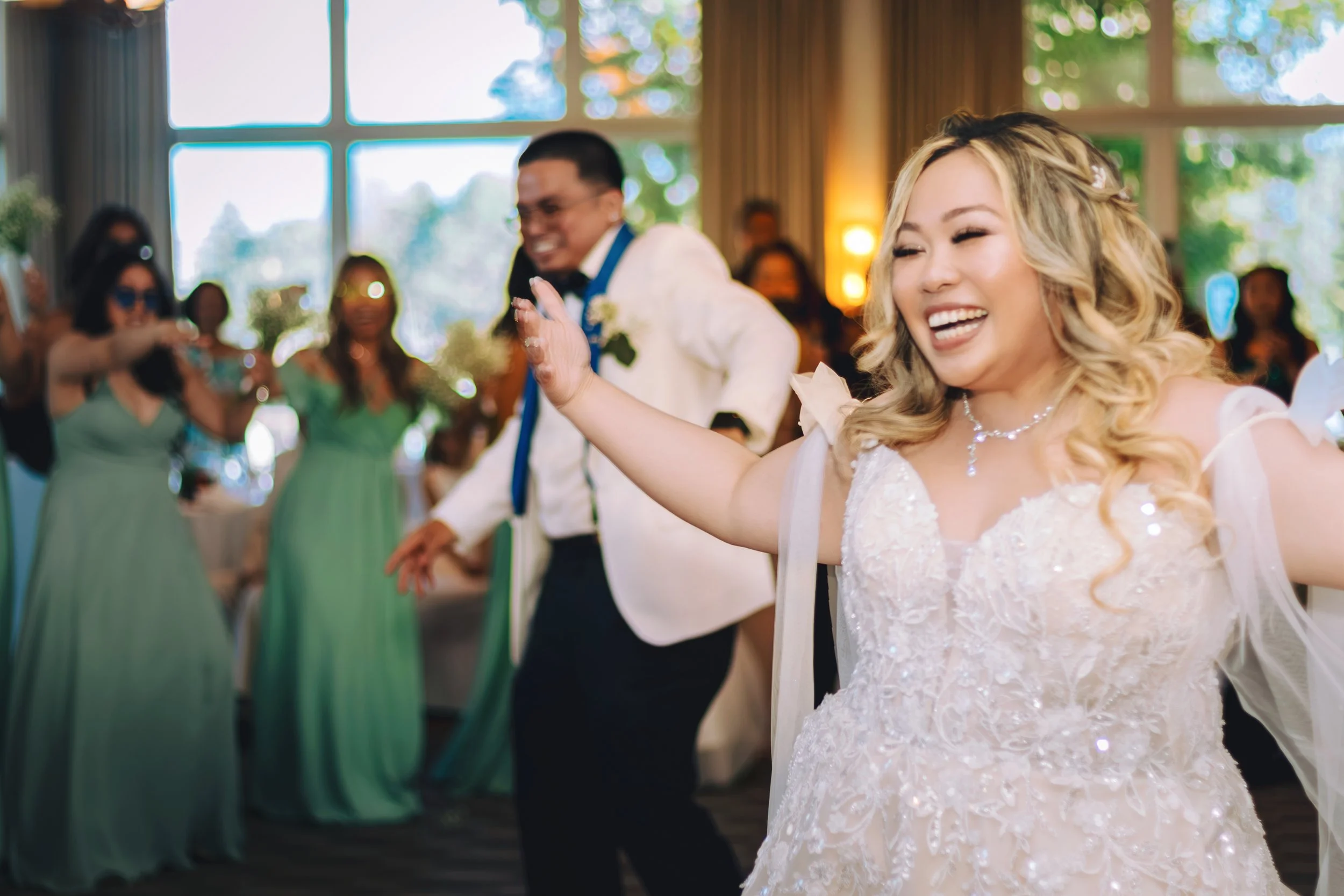 A bride and groom dancing joyfully at their wedding reception, surrounded by wedding guests, in a bright, elegant venue with large windows and green scenery outside.