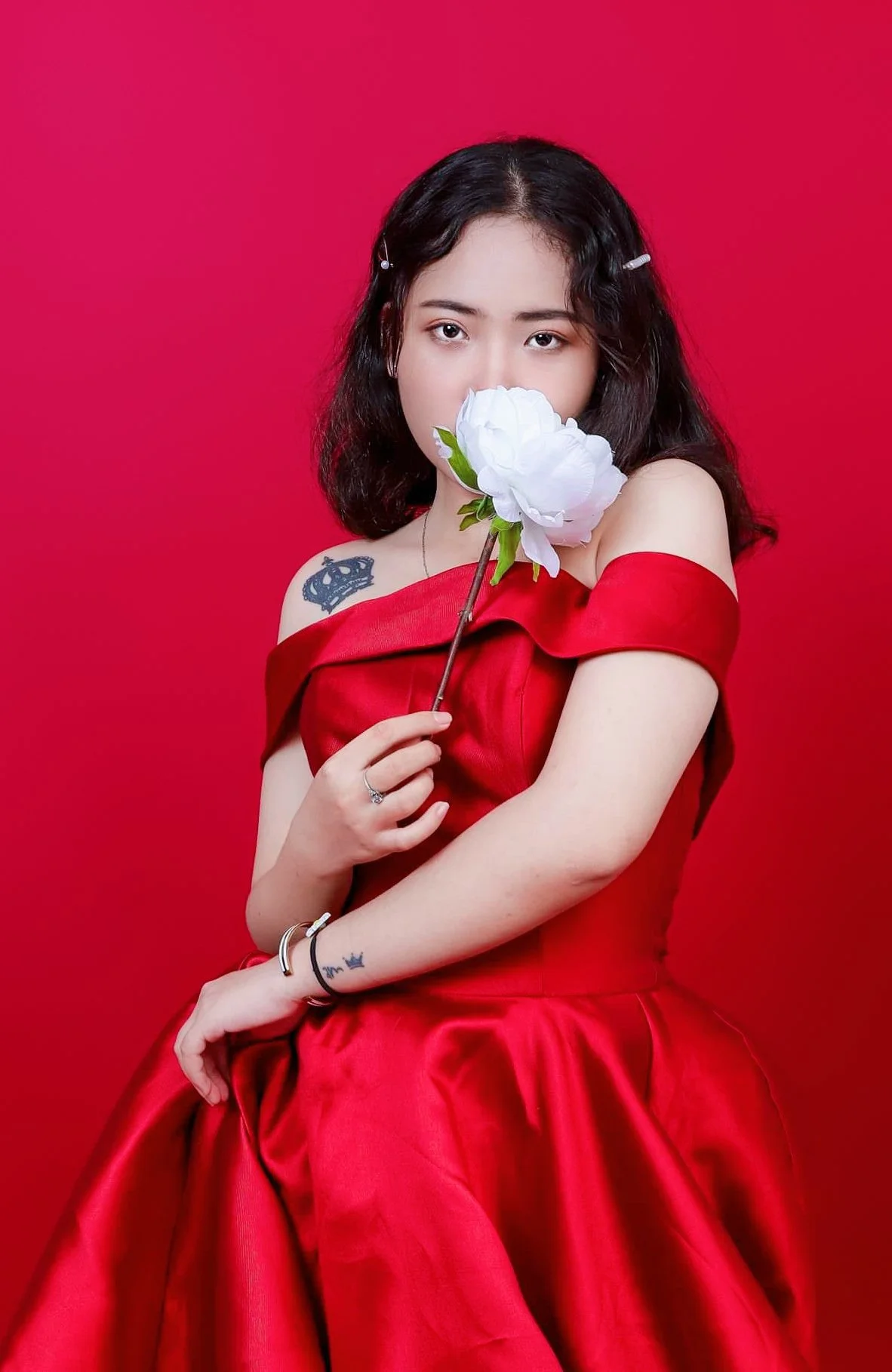 A woman with black hair, wearing a red off-shoulder dress, holding a large white flower near her face, against a red background.