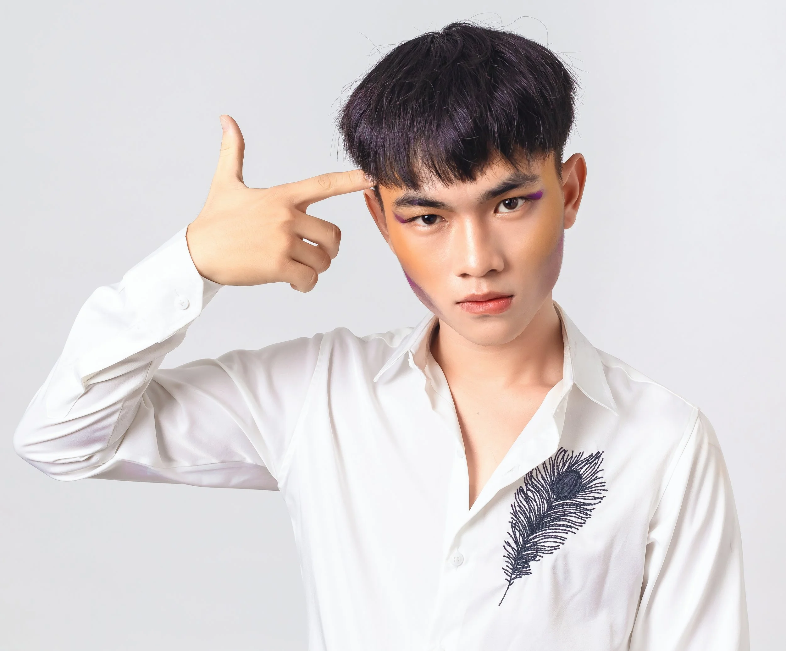 A young man with dark hair wearing a white shirt with a feather embroidered on it, raising his right hand to his head with a finger pointing to his temple, posing against a plain white background.