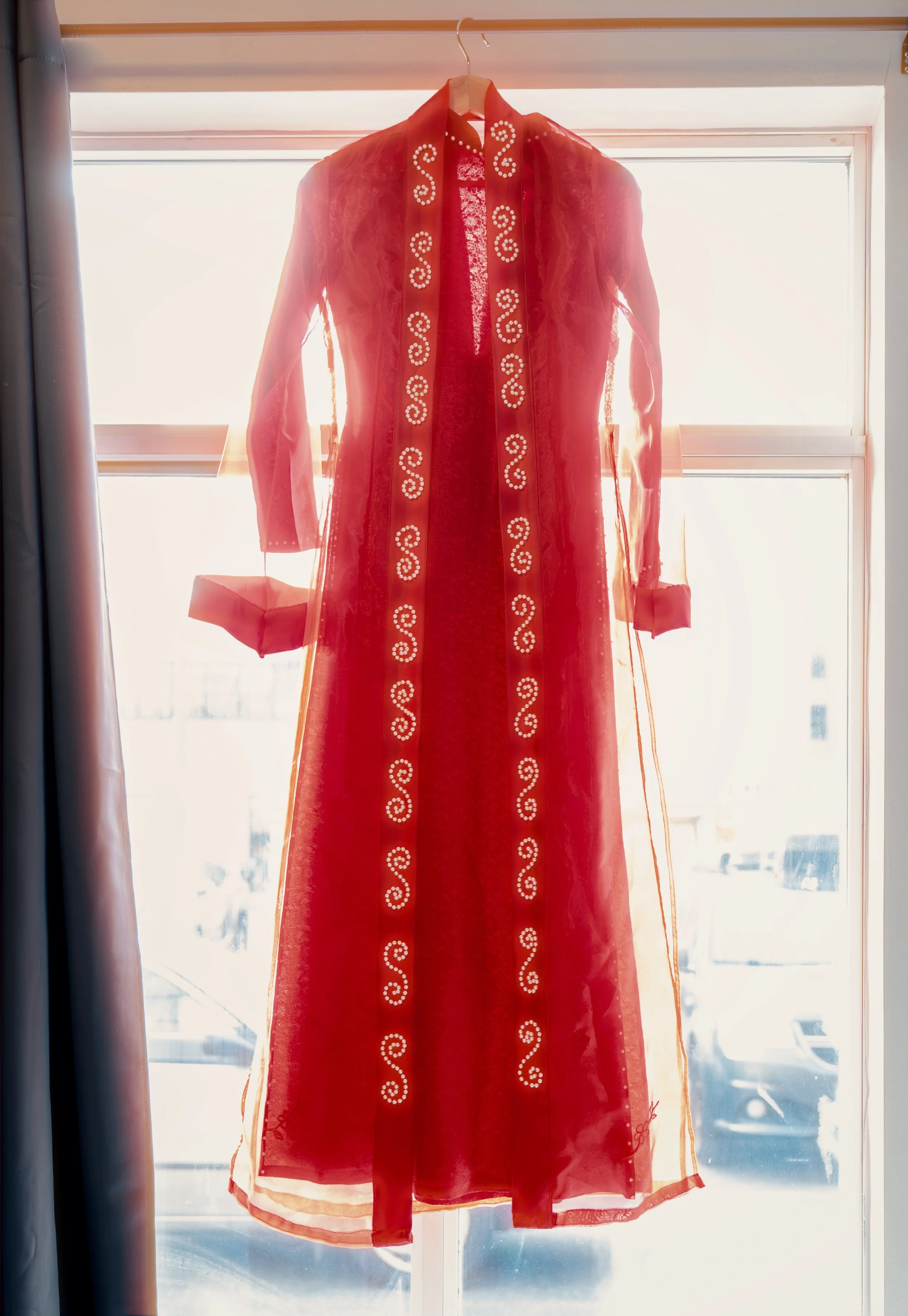 A red sheer dress with long sleeves and decorative white swirling embroidery hanging on a hanger in front of a window with sunlight shining through.