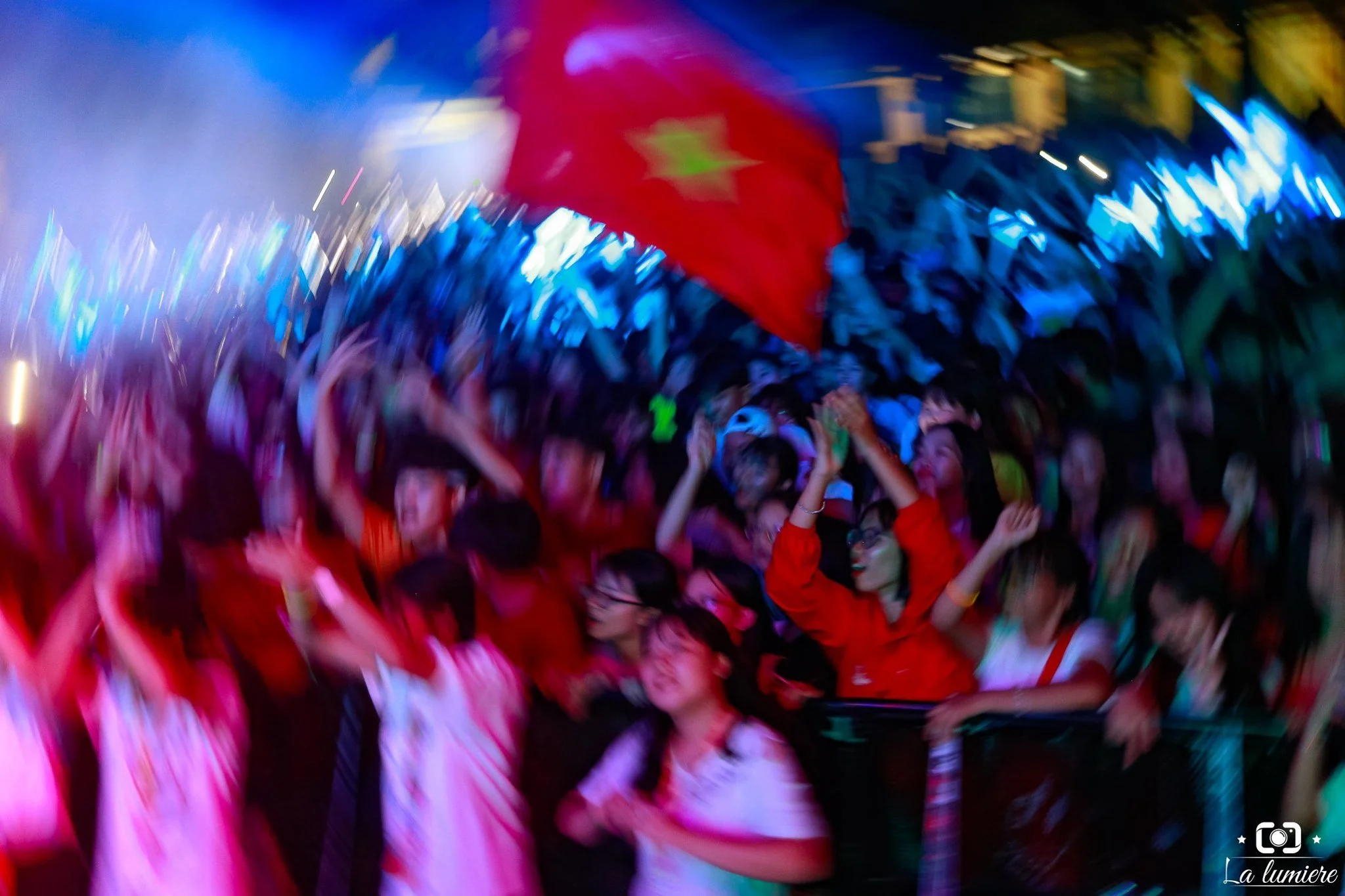 Blurred photo of a lively crowd at a concert or festival, with many people dancing and celebrating, holding a large red flag with a star.