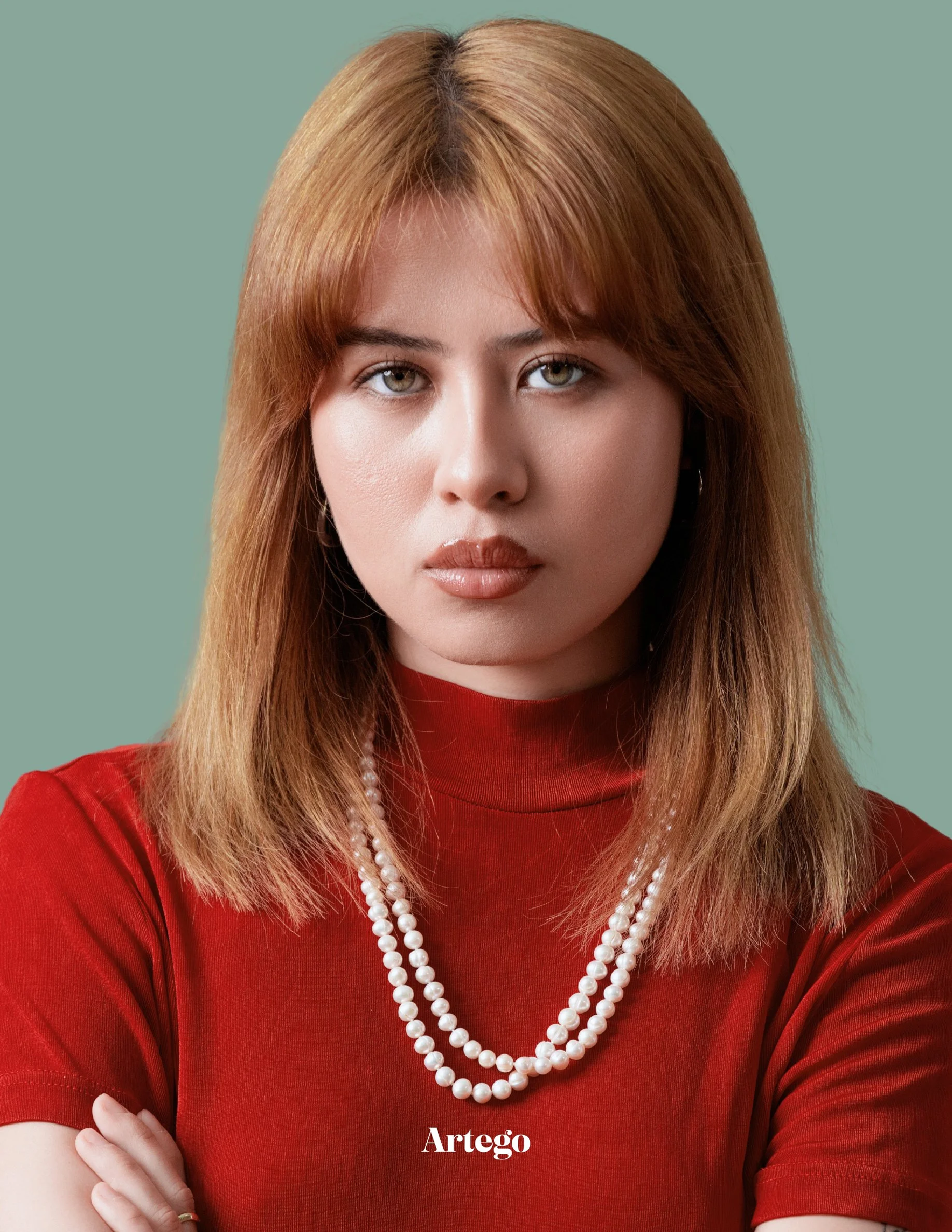 A woman with shoulder-length reddish-brown hair looking directly at the camera against a muted green background, wearing a red turtleneck and a string of pearls.