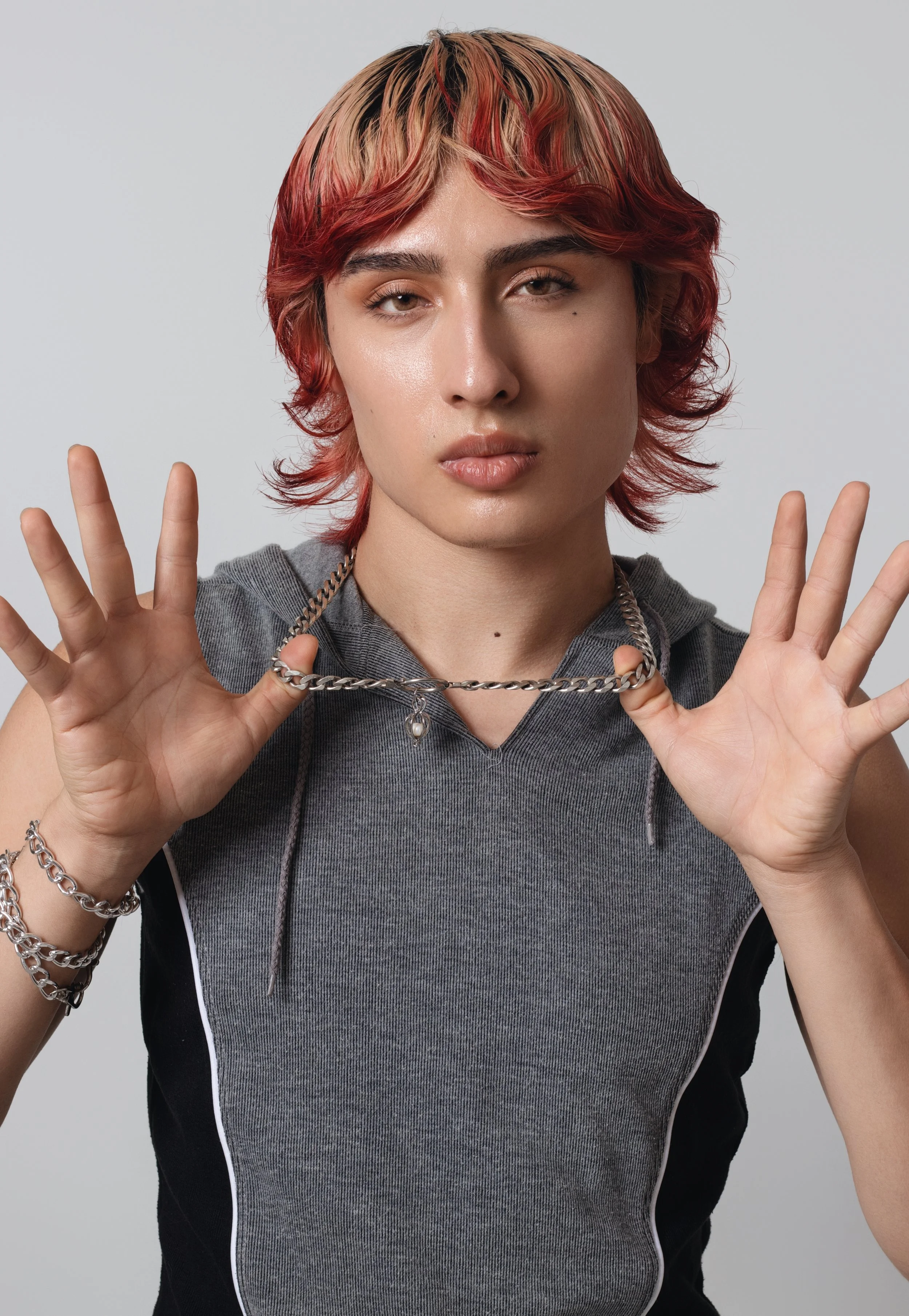 Person with short, layered hair dyed blonde and red, wearing a sleeveless gray and black athletic top, holding a silver chain necklace with their hands.