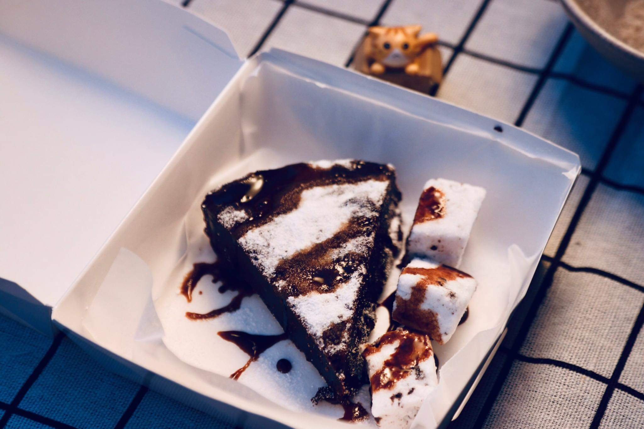 A slice of chocolate cake with powdered sugar, served with marshmallows topped with chocolate syrup in a white takeout box.