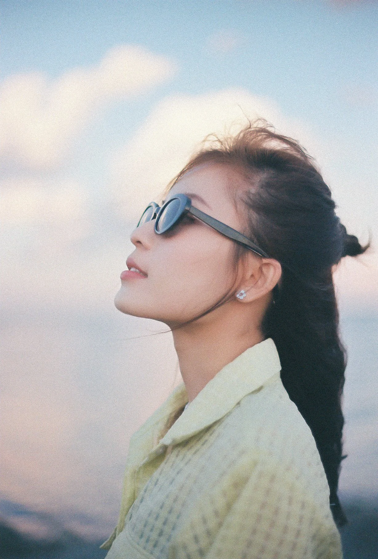 Profile of a woman with long dark hair, wearing sunglasses and earrings, outdoors with a cloudy sky background.