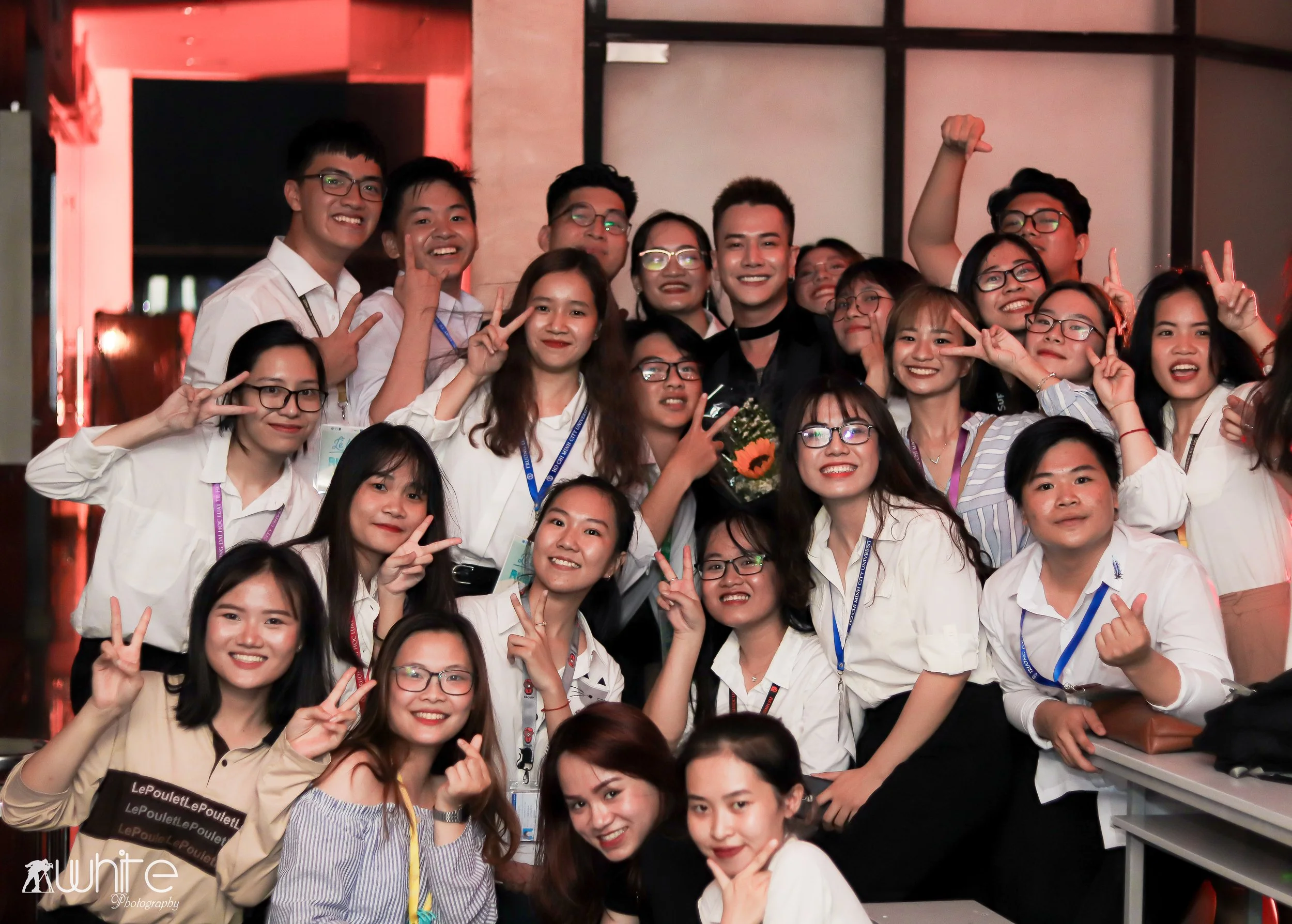 Group of young people smiling, posing, and making peace signs at an indoor event, some wearing glasses and conference badges, with a woman in the center holding a flower bouquet.